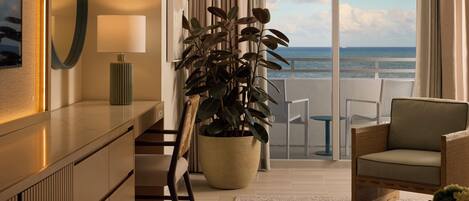 A hotel room with a large potted plant, a chair, a table, a lamp, and a view of the ocean through large windows.