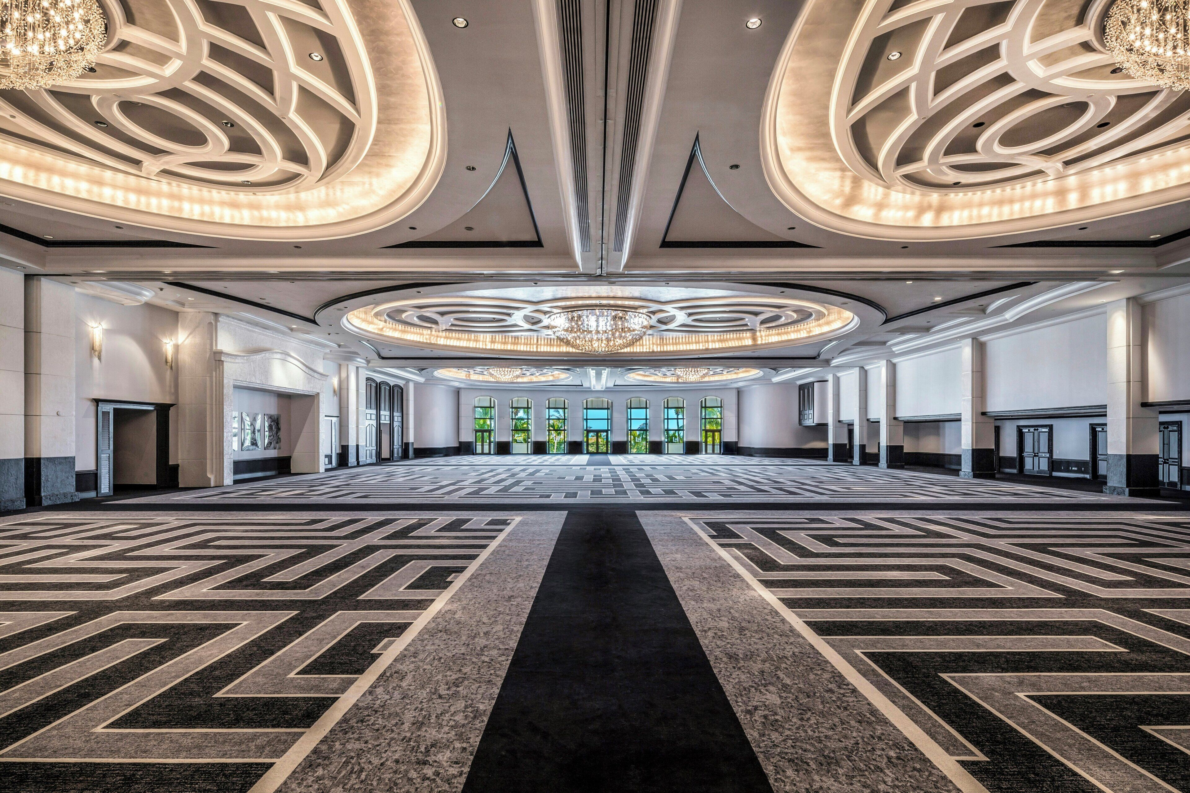 A spacious hotel lobby with a central chandelier, patterned carpet, and symmetrical wall design.