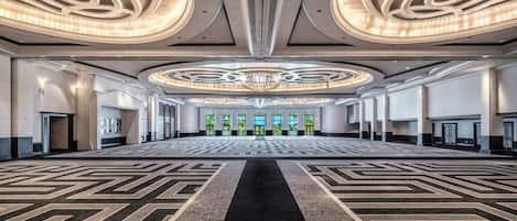 A spacious hotel lobby with a central chandelier, patterned carpet, and symmetrical wall design.