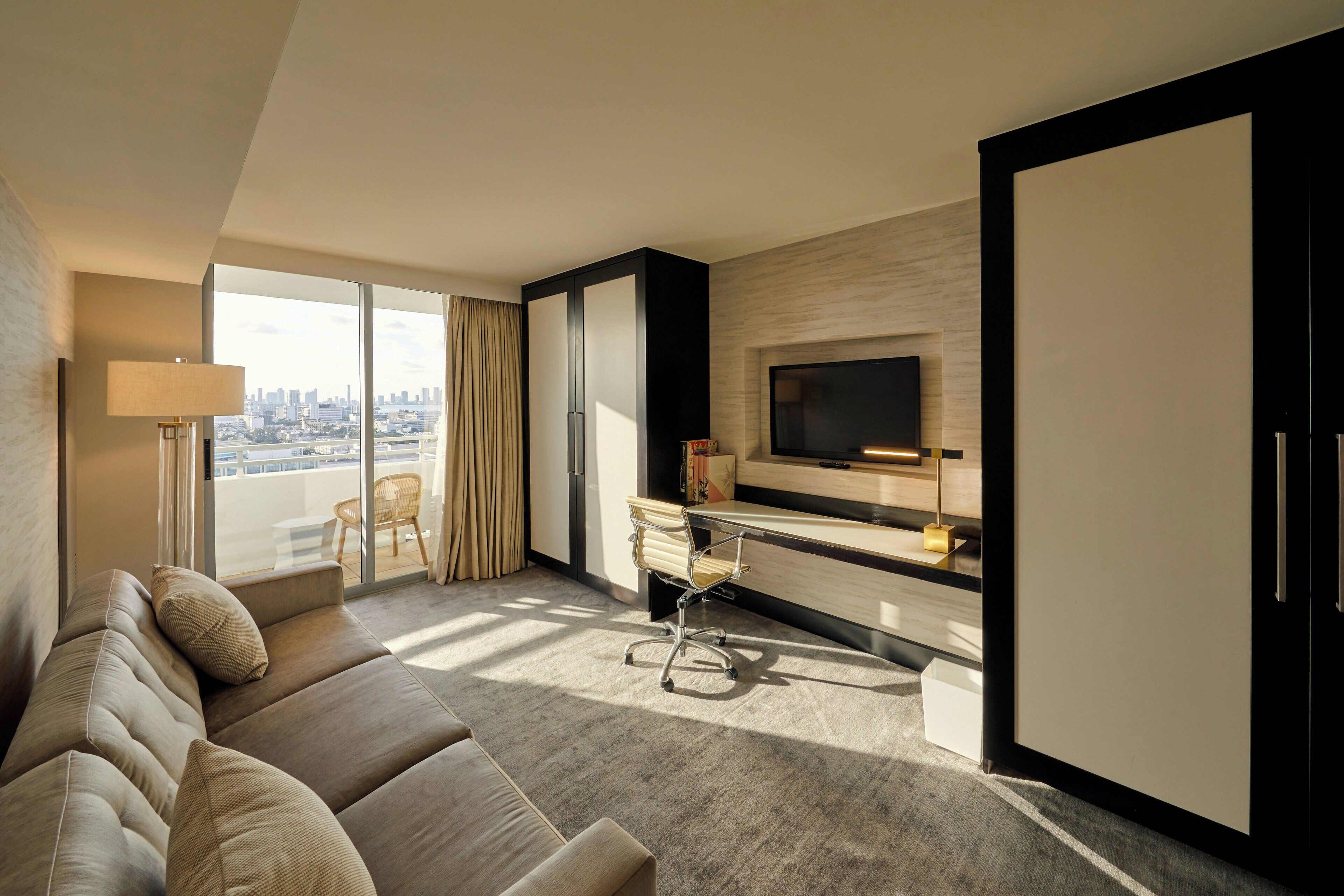 A modern hotel room with a large sofa, a desk, a TV, and a city view.
