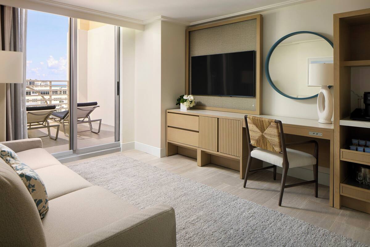 Une chambre d’hôtel avec un canapé, un bureau avec un ordinateur, une chaise, une télévision, un miroir et une vue sur la ville.