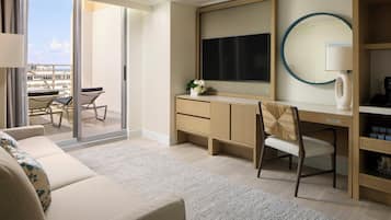 Quarto de hotel com sofá, escrivaninha com computador, cadeira, televisão, espelho e vista para a cidade.