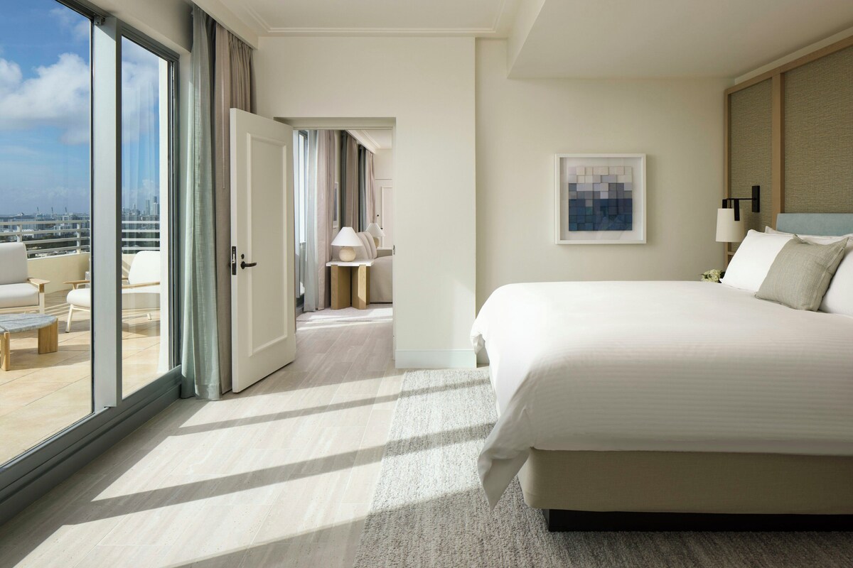 Une chambre d’hôtel avec un grand lit, une vue dégagée sur la ville grâce à une porte coulissante et un panorama urbain visible à l’extérieur.