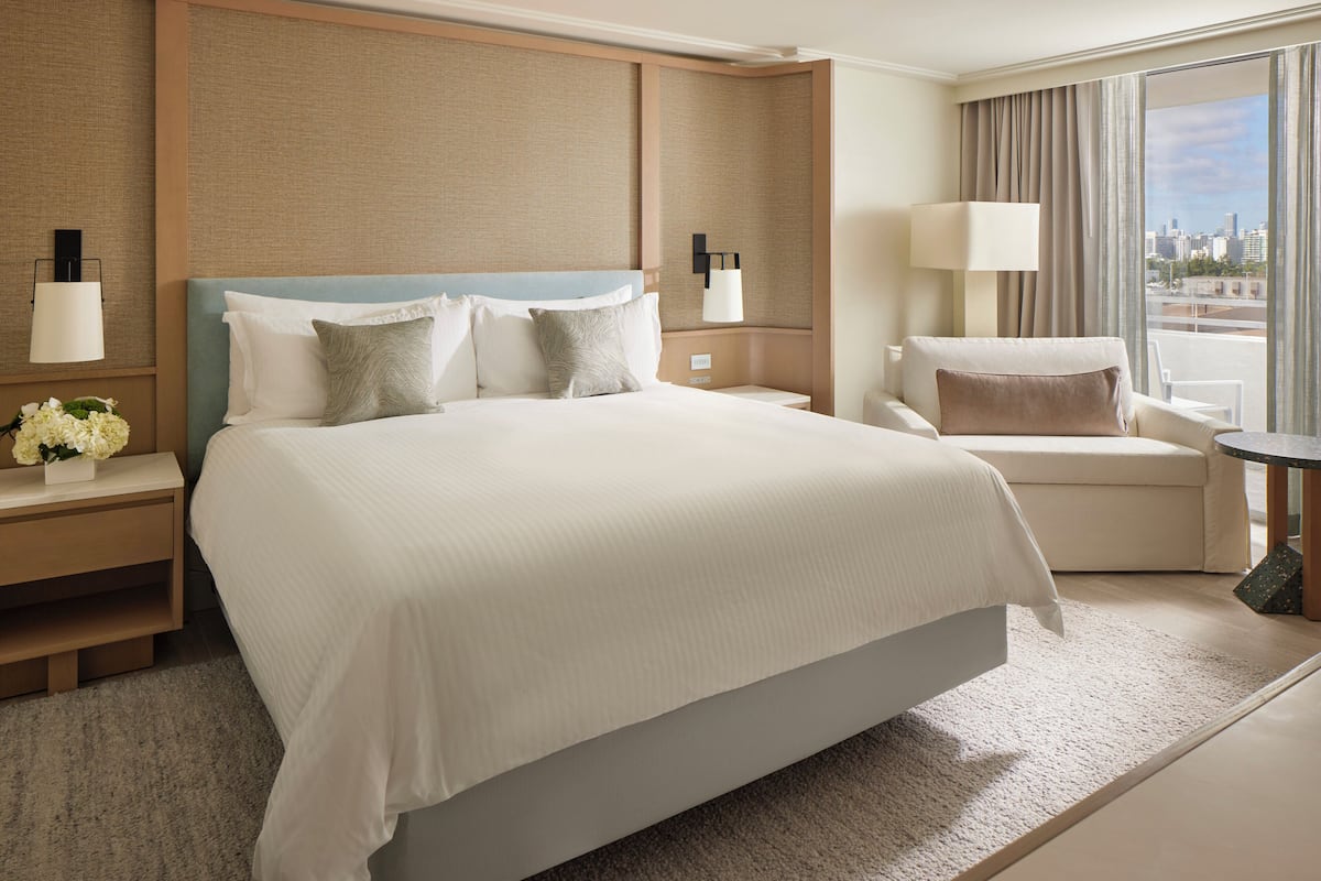 Une chambre d’hôtel avec un grand lit, des tables de chevet avec des lampes, une chaise, une petite table et une vue sur la ville.