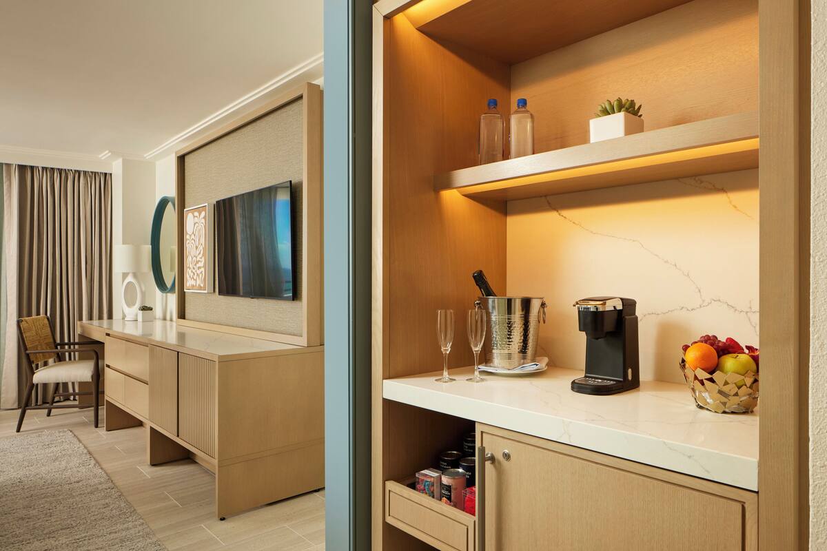 Une chambre d’hôtel moderne avec un bureau en bois, une machine à café et un panier de fruits.