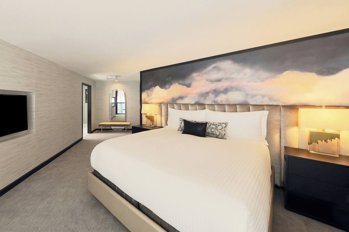 Une chambre d’hôtel moderne dotée d’un grand lit, d’un téléviseur à écran plat fixé au mur, d’une grande fenêtre et d’un tableau représentant des nuages.