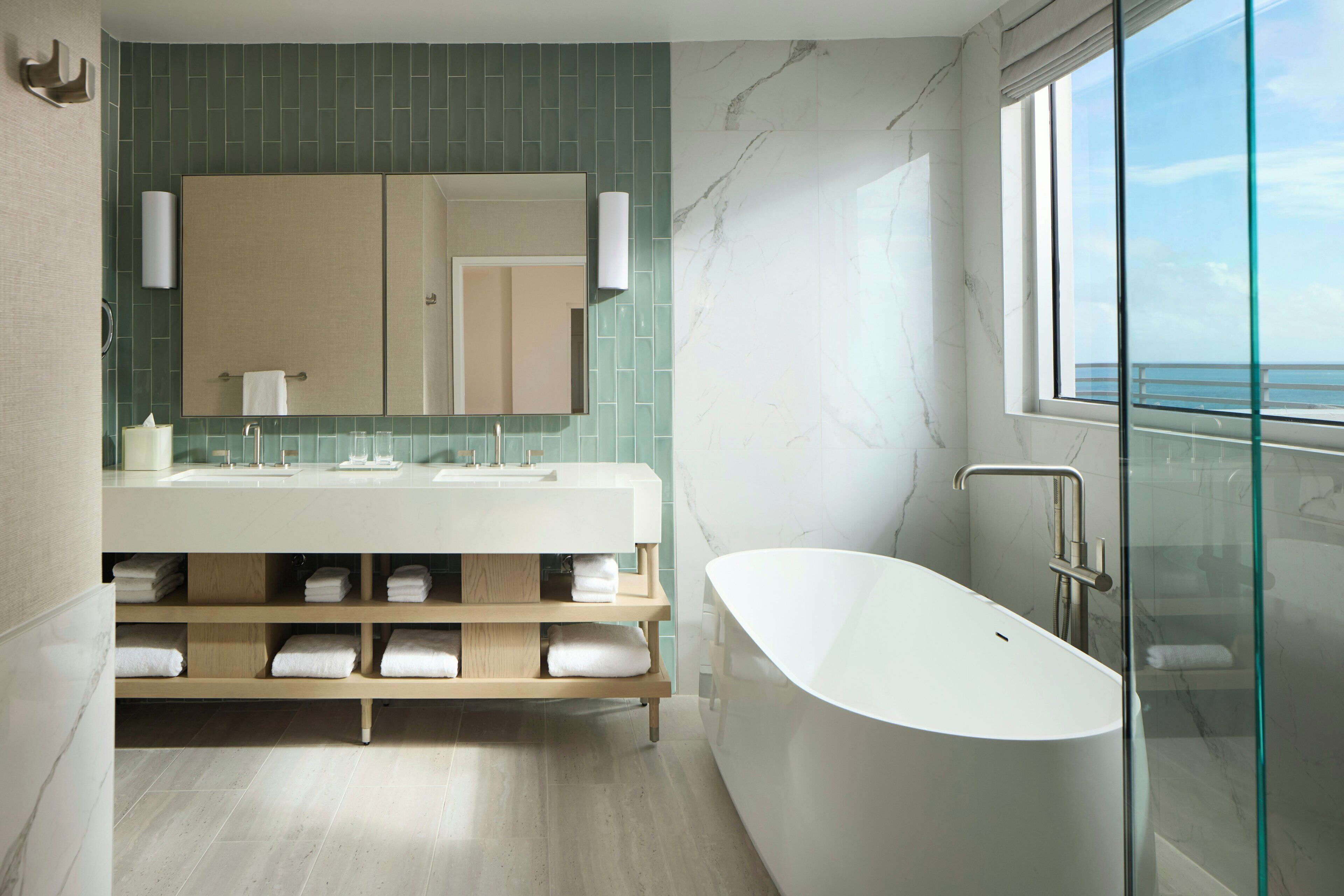 Une salle de bain moderne avec une baignoire sur pieds, un grand miroir et une vue sur la mer grâce à une porte en verre.
