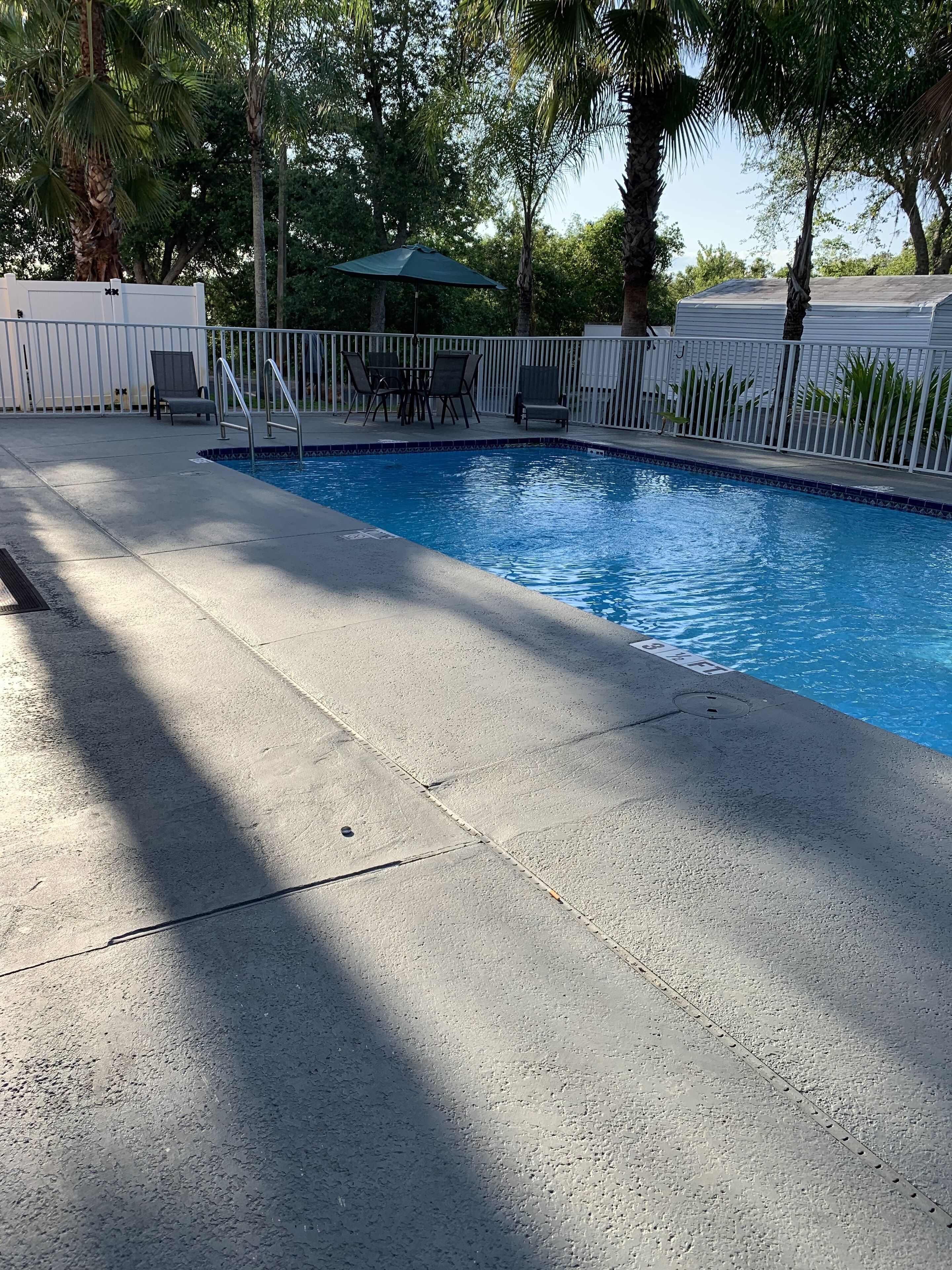 A swimming pool with a tiled edge, a covered seating area with chairs, and a white fence surrounding the pool.