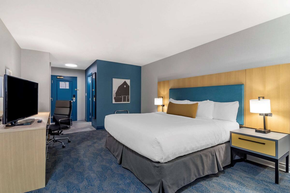 A hotel room with a bed, bedside table, lamp, desk with a TV, and a chair.