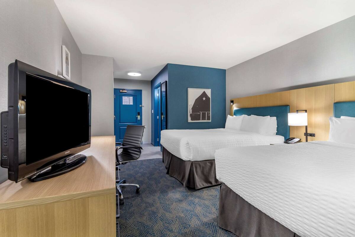 A hotel room with a TV, two beds, a desk, a chair, and a blue wall with a picture.
