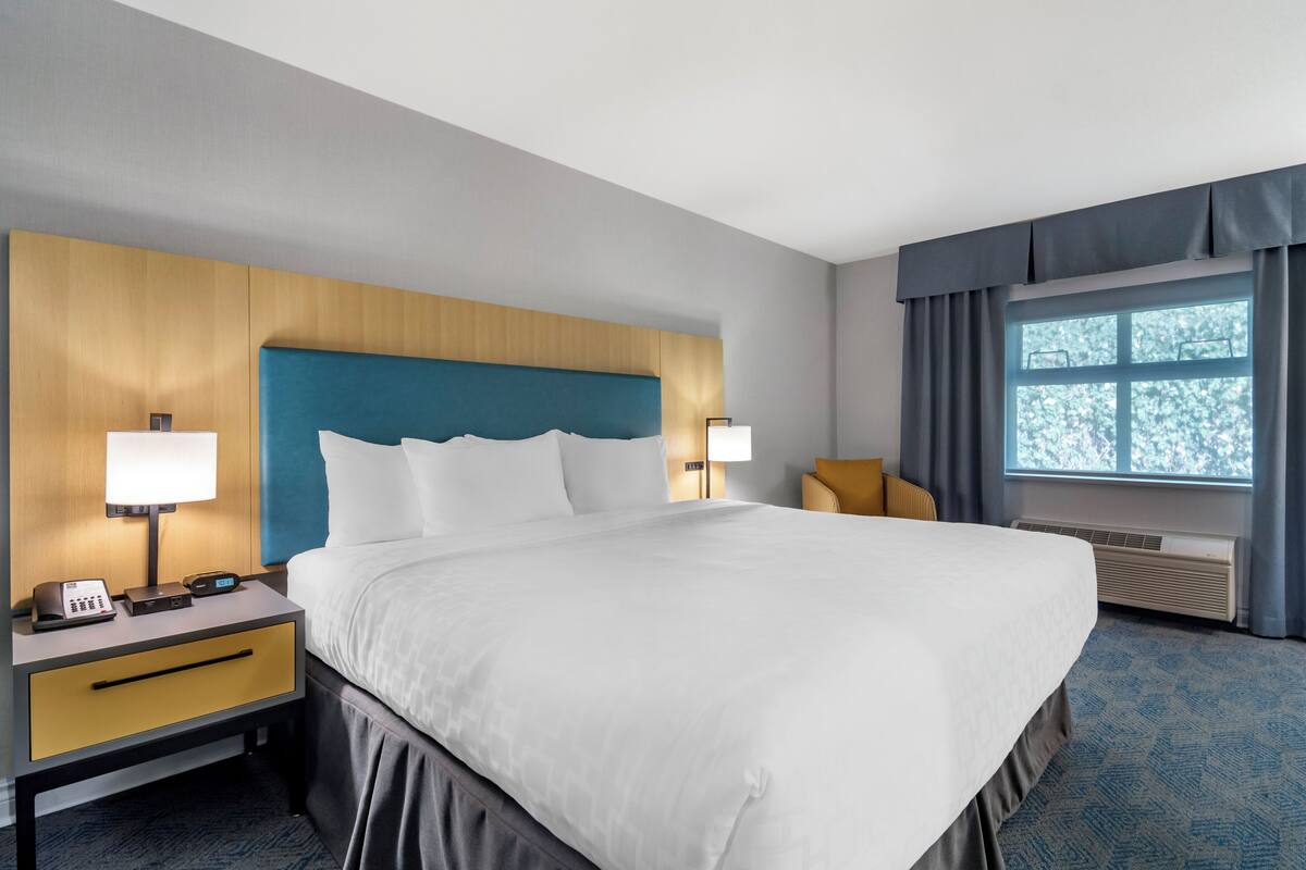 A hotel room with a large bed, two bedside tables with lamps, a window with curtains, and a wall-mounted air conditioning unit.