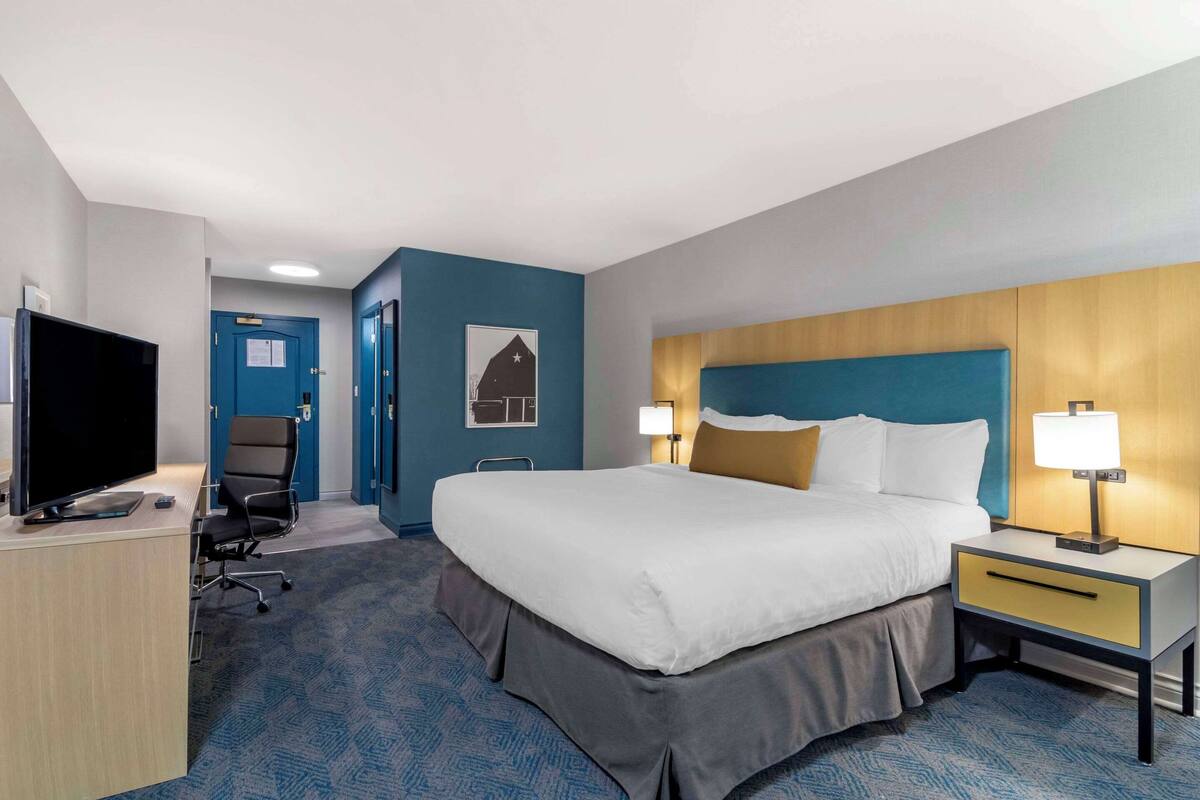 A hotel room with a bed, bedside table, lamp, TV, desk, chair, and a door.