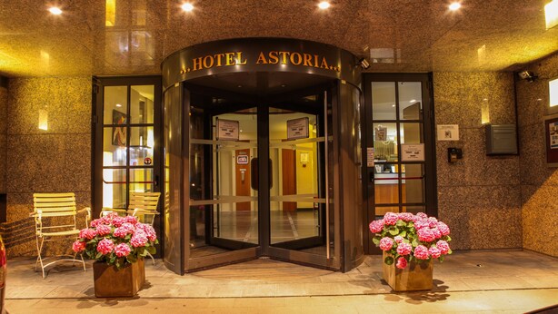 La entrada del Hotel Astoria, con una puerta giratoria, dos plantas en macetas con flores rosadas y bancos de madera en el exterior.