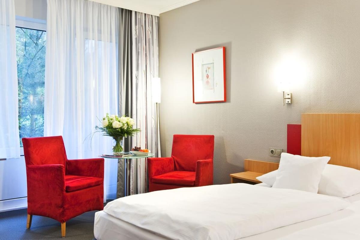 Une chambre d’hôtel avec un lit, deux fauteuils rouges, une petite table sur laquelle est posé un vase de fleurs, un tableau encadré au mur et une fenêtre avec des rideaux.