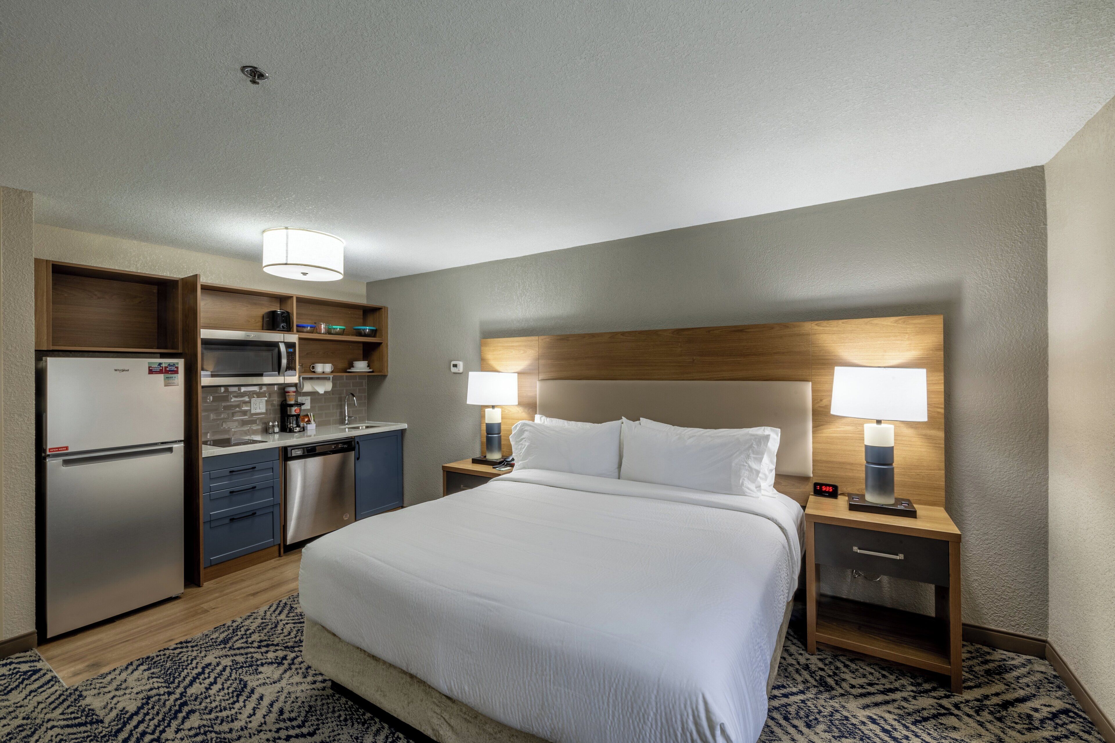 A hotel room with a bed, two bedside tables with lamps, a built-in desk, a microwave, a refrigerator, and a coffee maker.