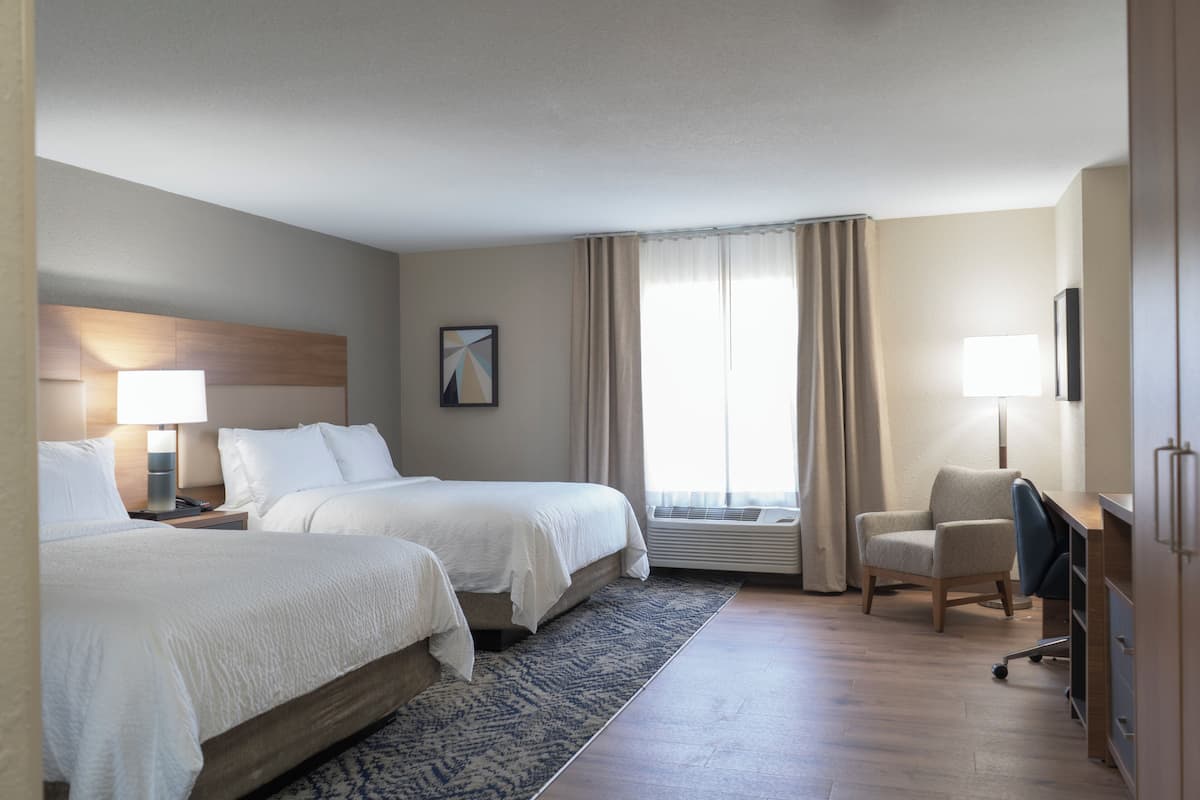 A hotel room with two beds, a desk, a chair, a lamp, a picture on the wall, and a window with curtains.