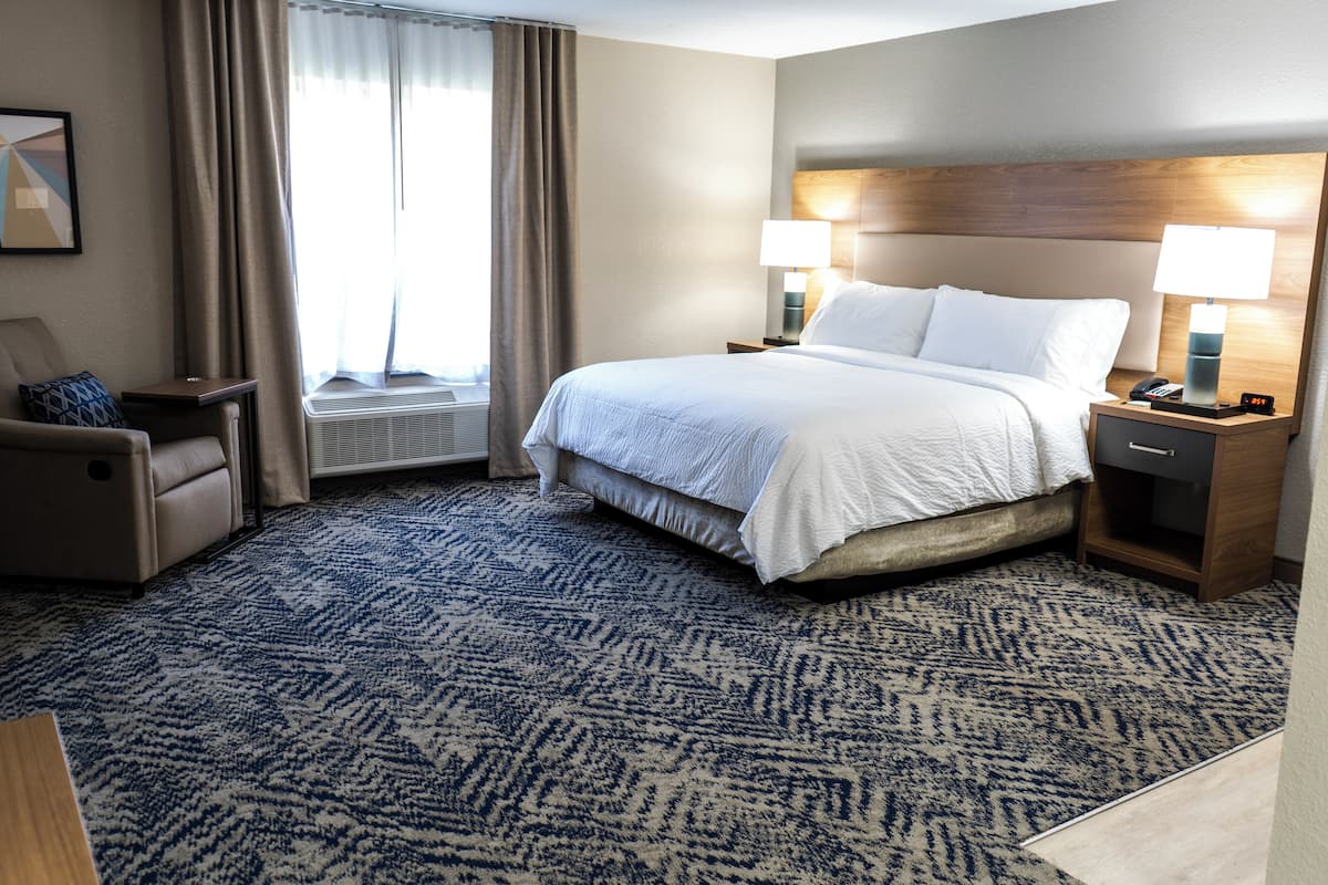 A hotel room with a large bed, two bedside tables with lamps, a chair, a window with curtains, and a patterned carpet.