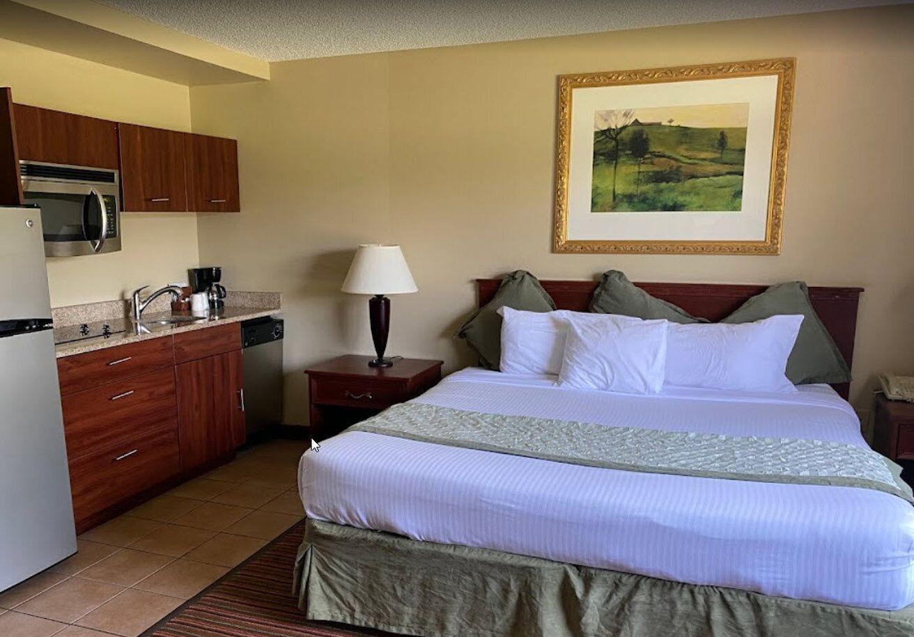 A hotel room with a bed, a nightstand, a lamp, a painting on the wall, and a kitchenette with a microwave and coffee maker.