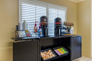 Una estación de café con varias cafeteras, un dispensador de desinfectante de manos y una bandeja de muffins.