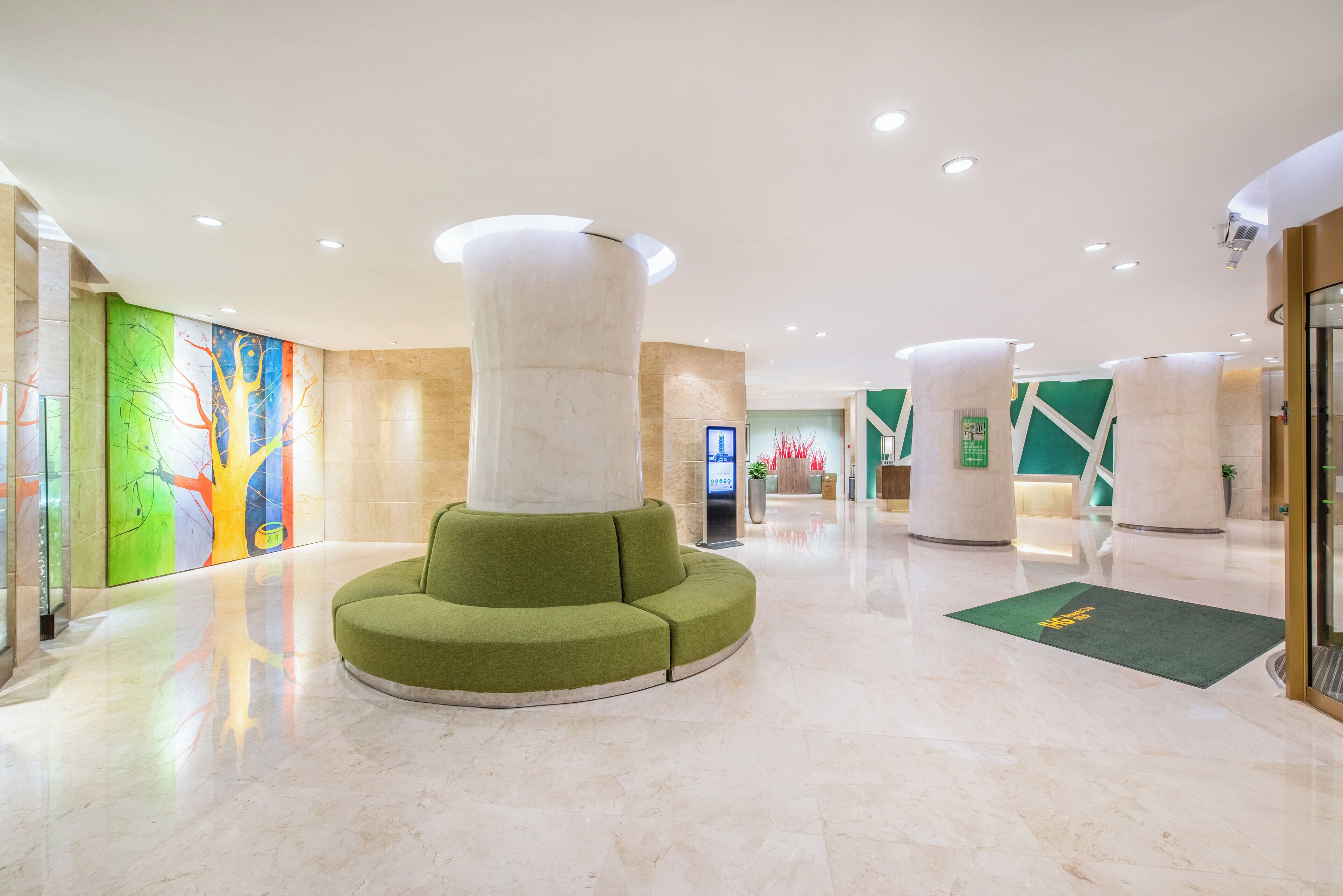 A modern lobby with a central green seating area, a digital information kiosk, and a glass elevator.