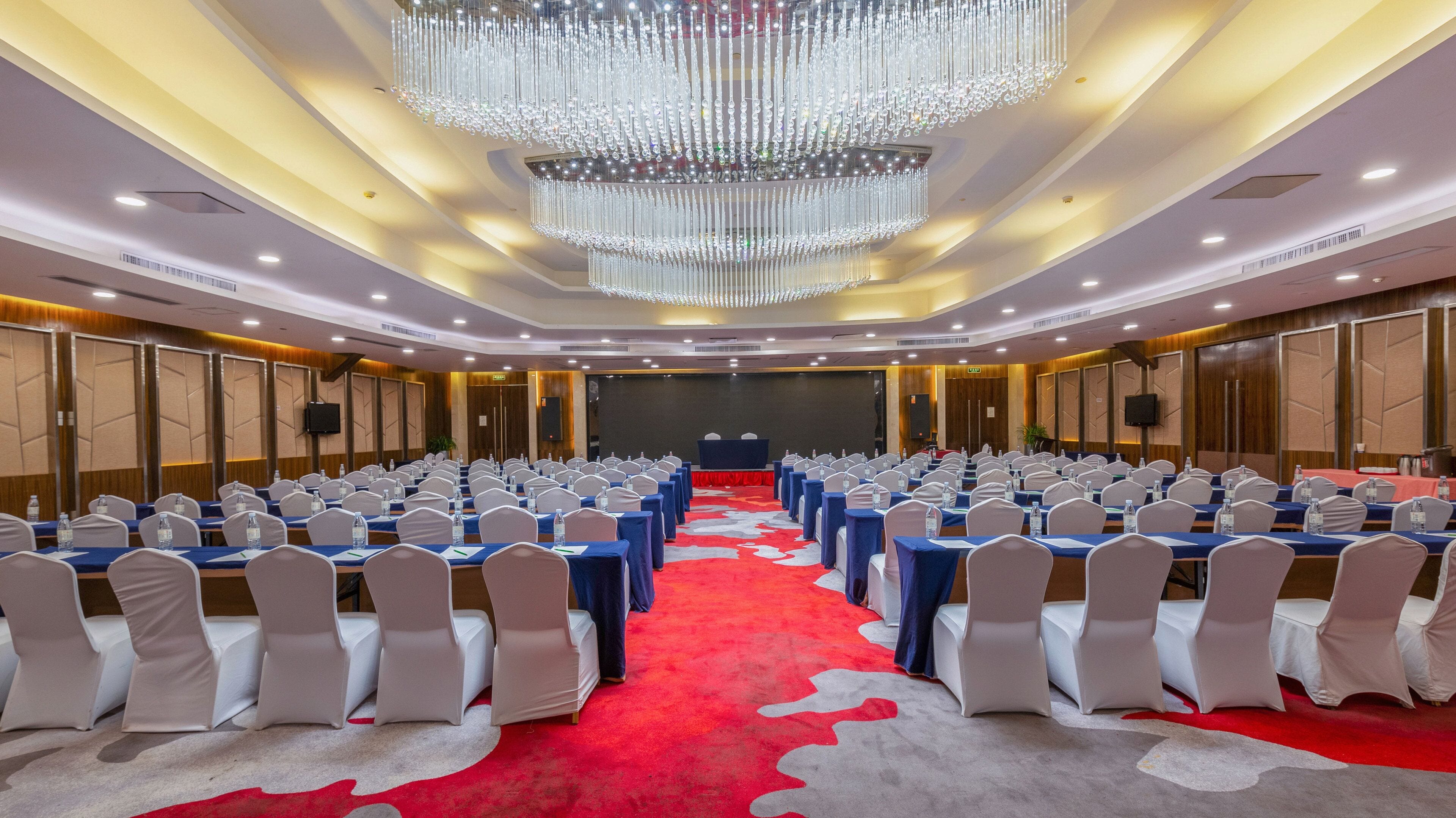 Un salón de conferencias con filas de mesas y sillas, una gran lámpara de araña y un escenario al frente.
