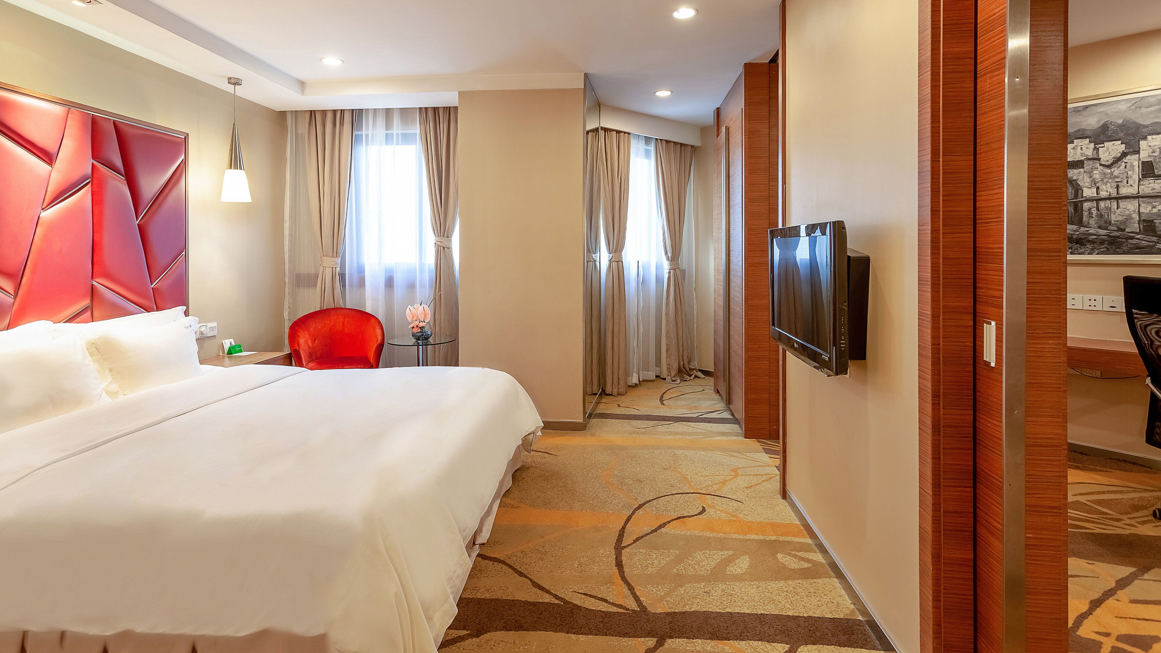A hotel room with a large bed, a red armchair, a small table with a vase, and a television mounted on the wall.