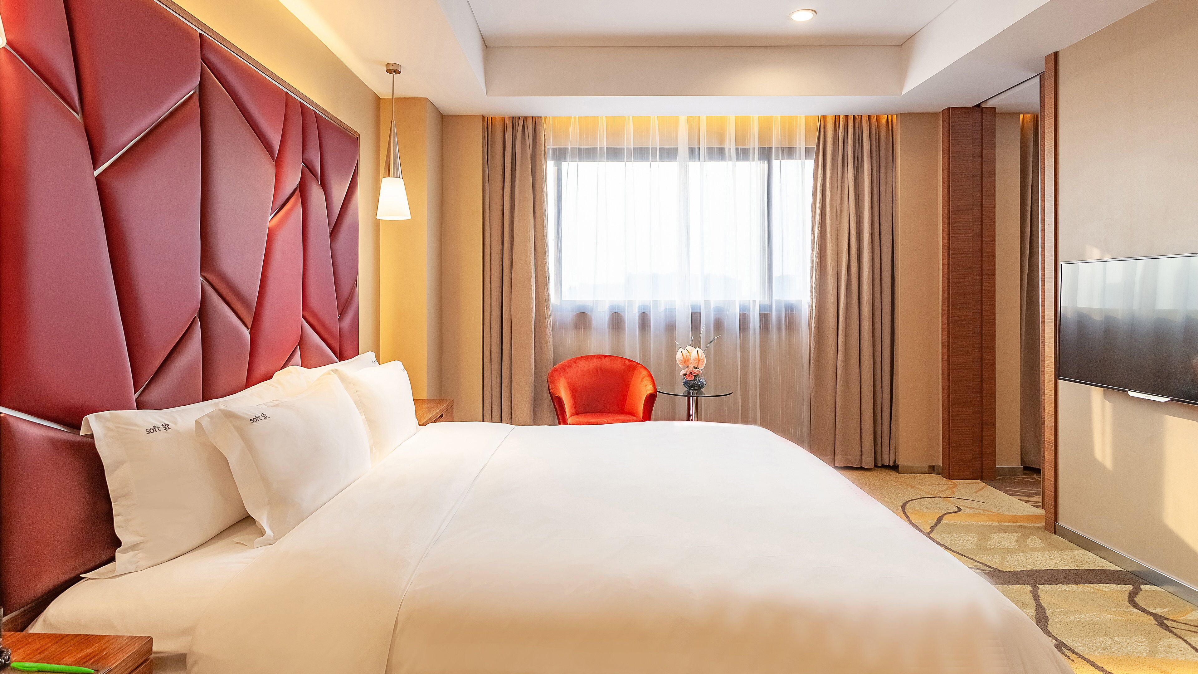 Habitación de hotel con una cama grande, un sillón rojo, una mesita con un florero y un televisor montado en la pared.