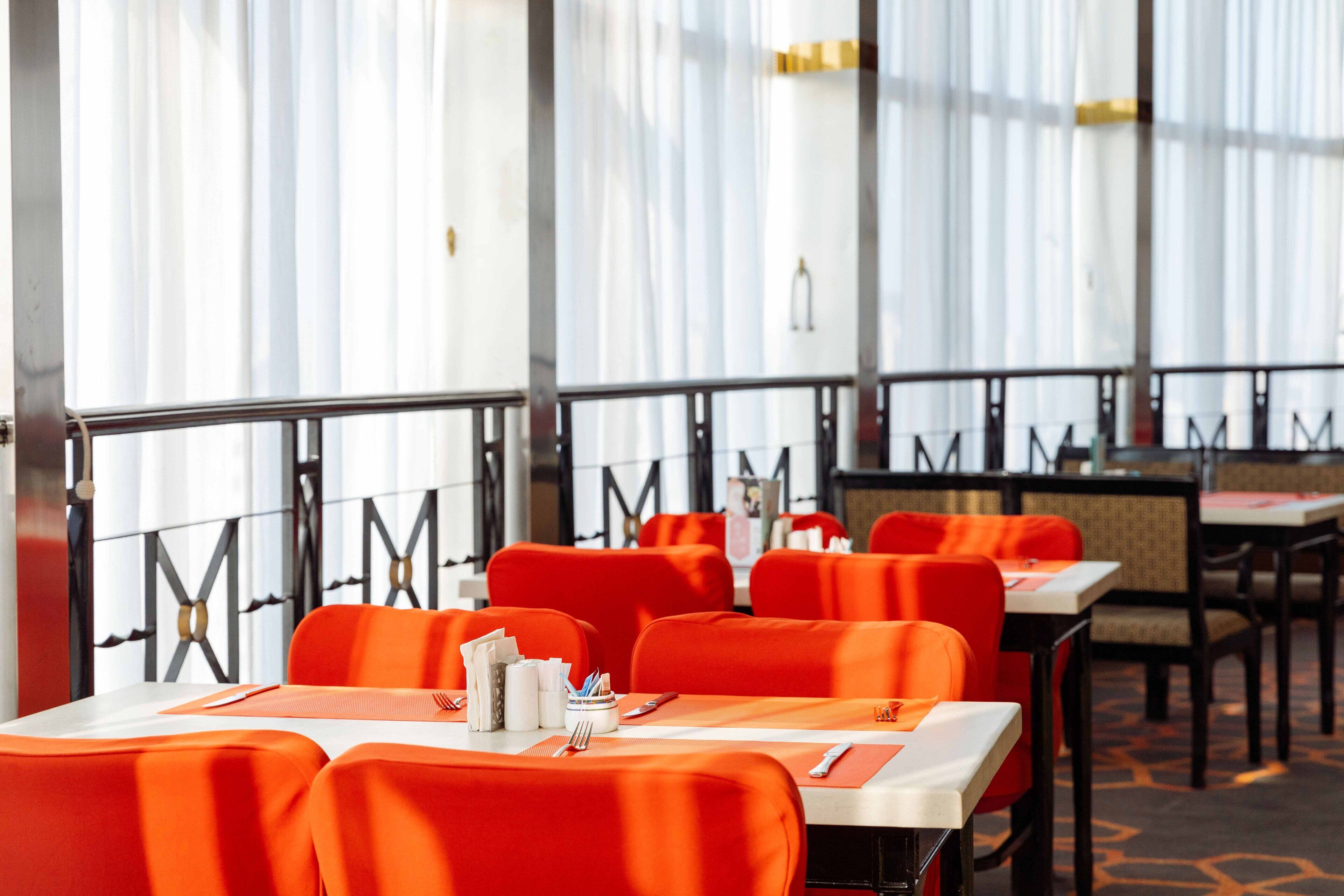 A restaurant with orange seating and white tablecloths, featuring a modern design with a glass railing and a view of the outside.