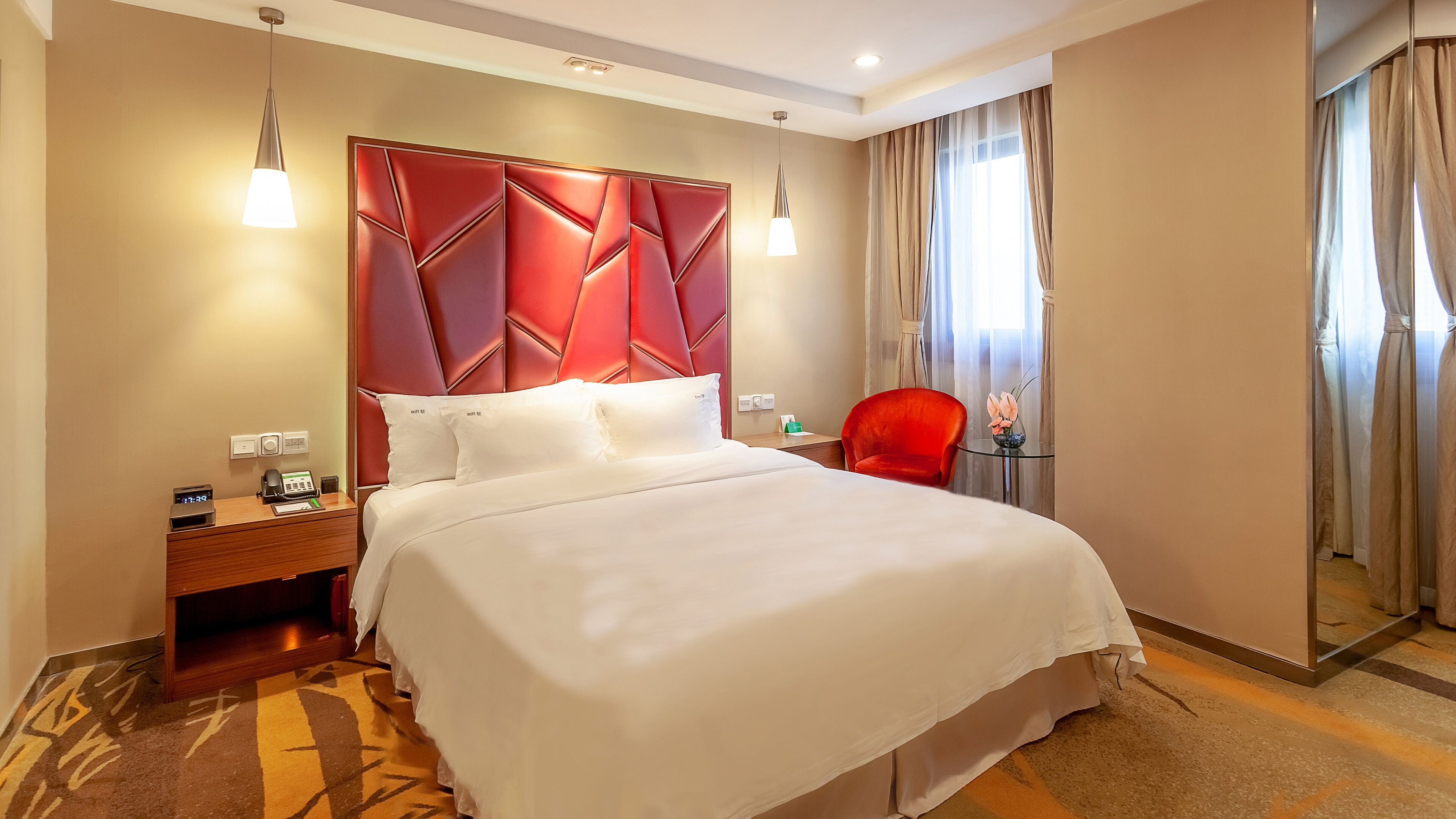 Habitación de hotel con una cama grande, cabecera tapizada de rojo, mesita de noche de madera, una silla roja, una pequeña mesa con un florero y un espejo en la pared.