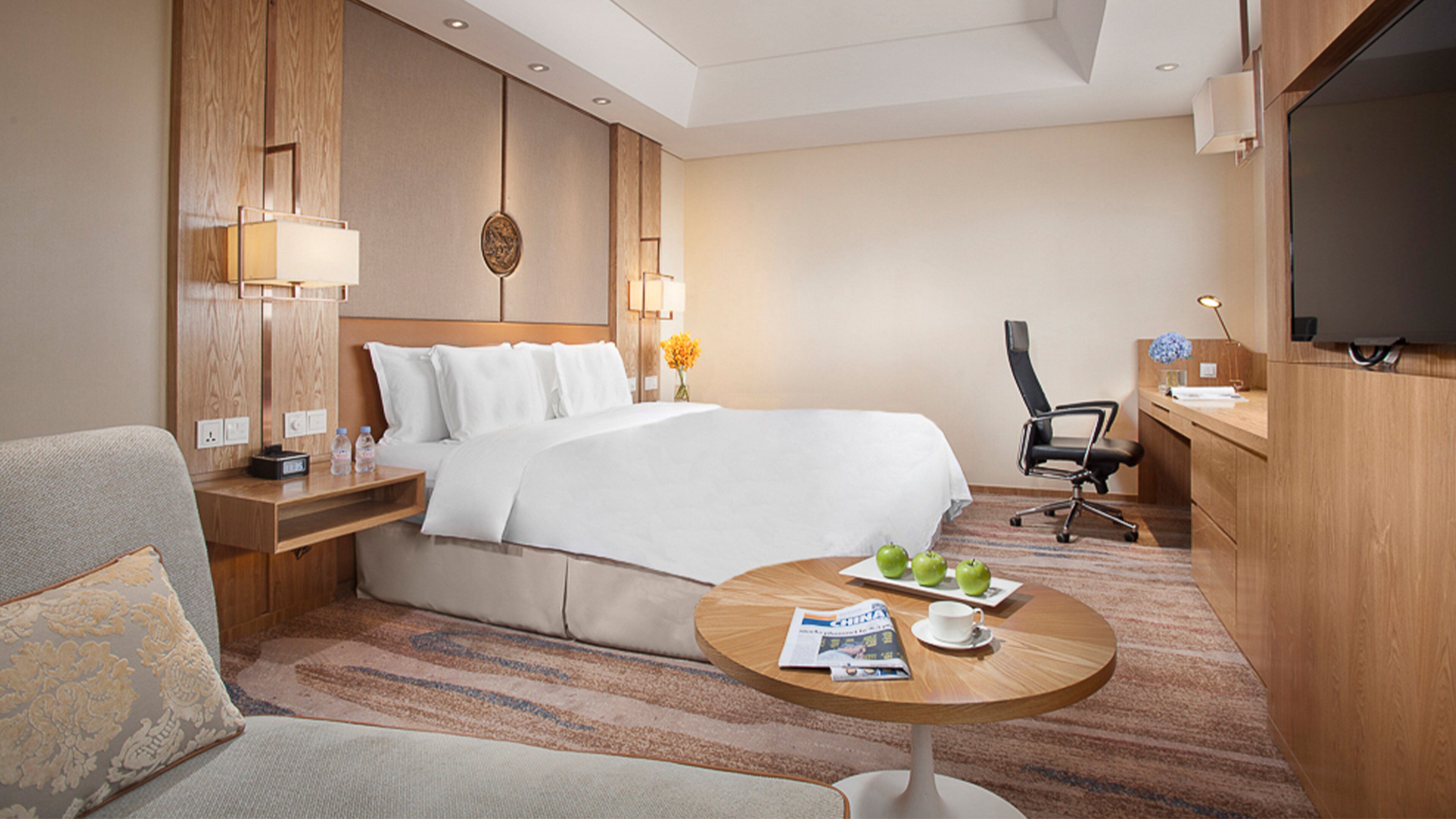 A hotel room with a bed, a desk, a chair, a television, and a round table with a magazine and a cup.