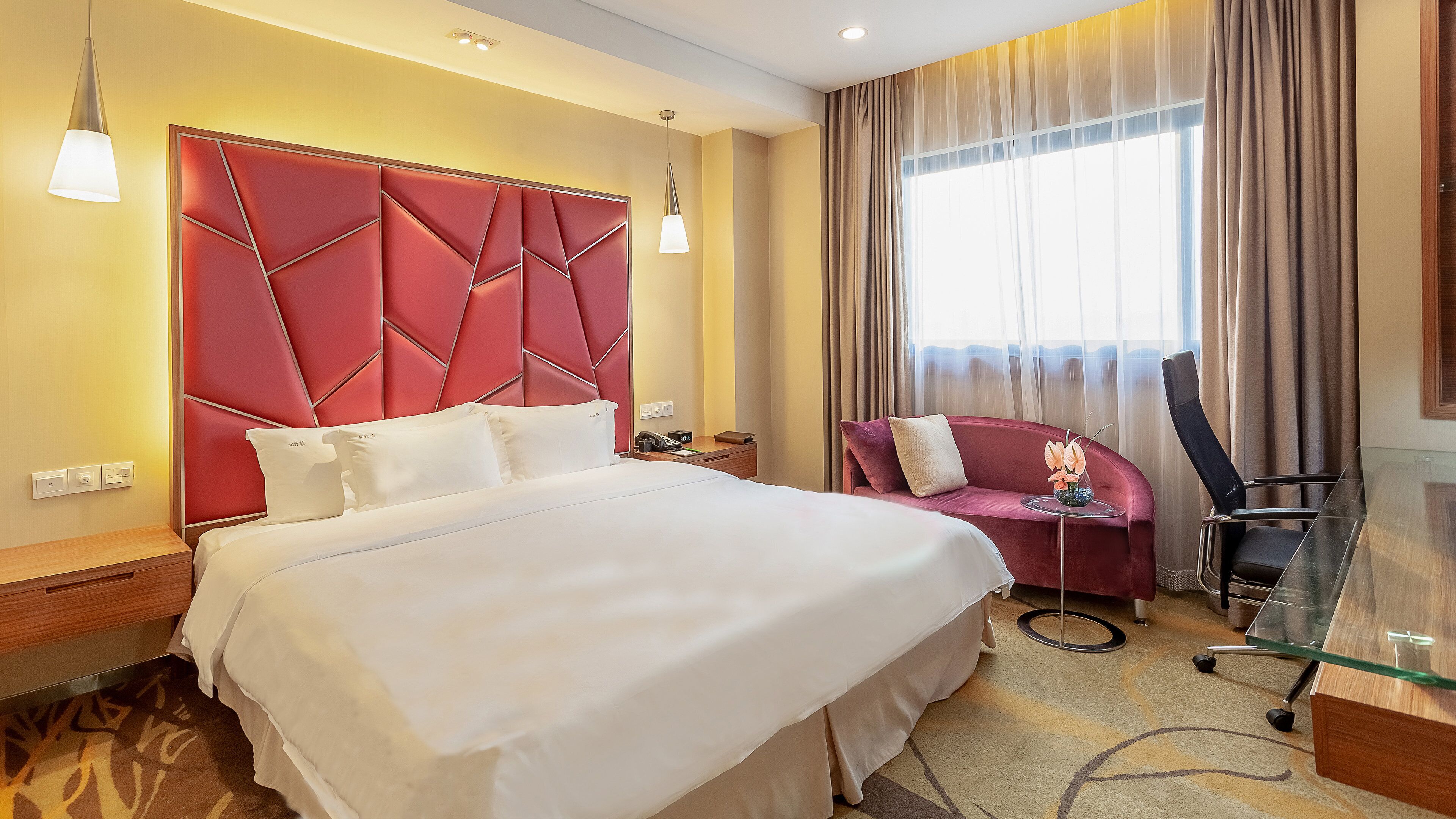 A hotel room with a large bed, a red headboard, a desk with a chair, and a large window with curtains.