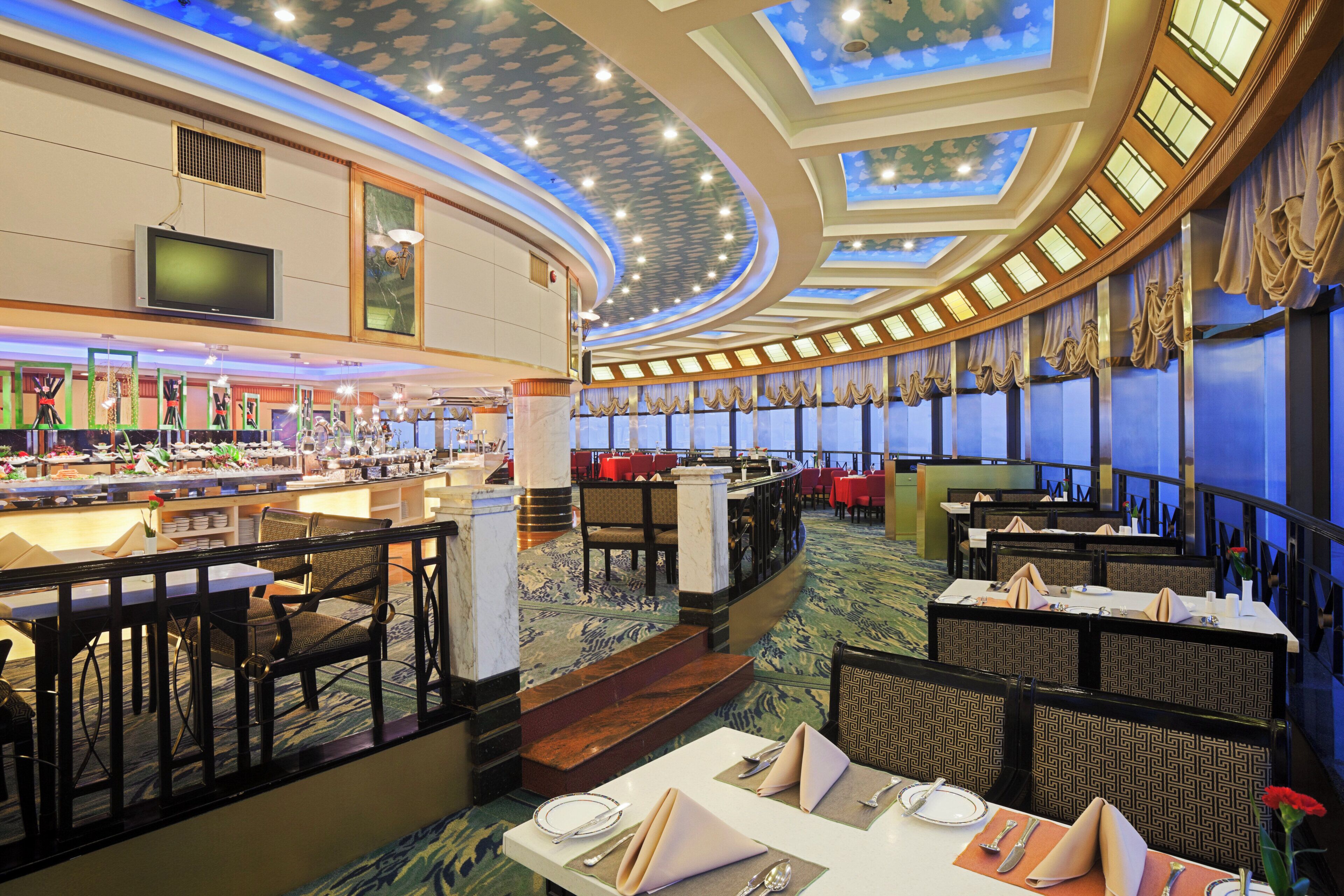 A hotel dining area with a central bar, multiple dining tables, and a ceiling with skylights.