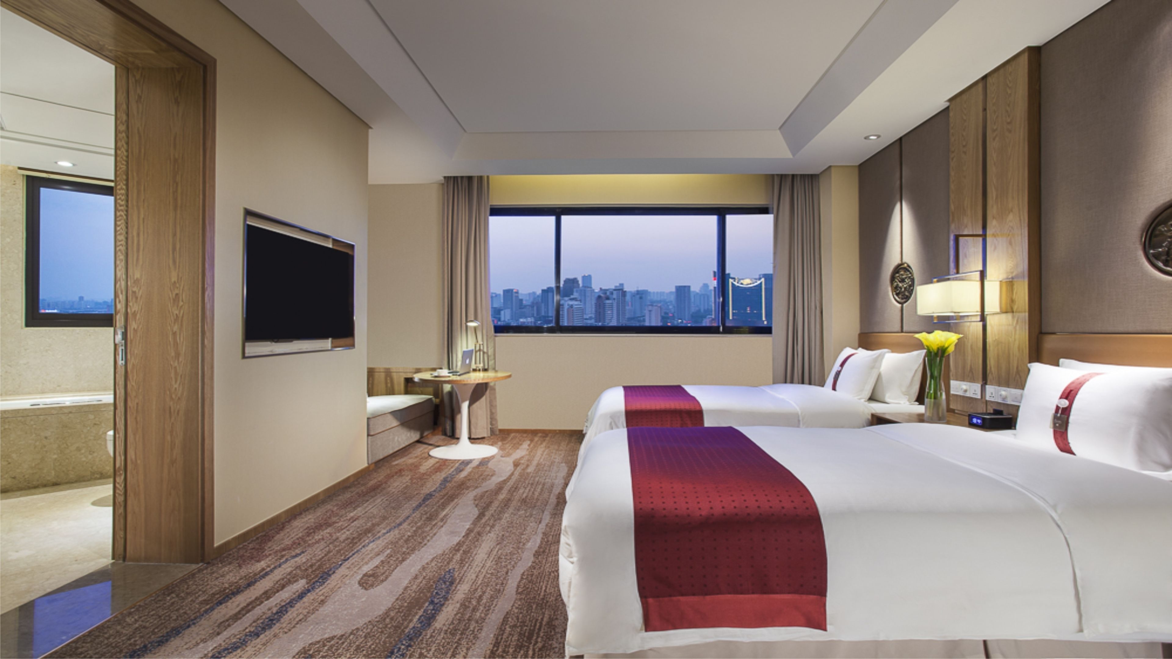 A hotel room with two beds, a TV, a desk, and a view of a cityscape.