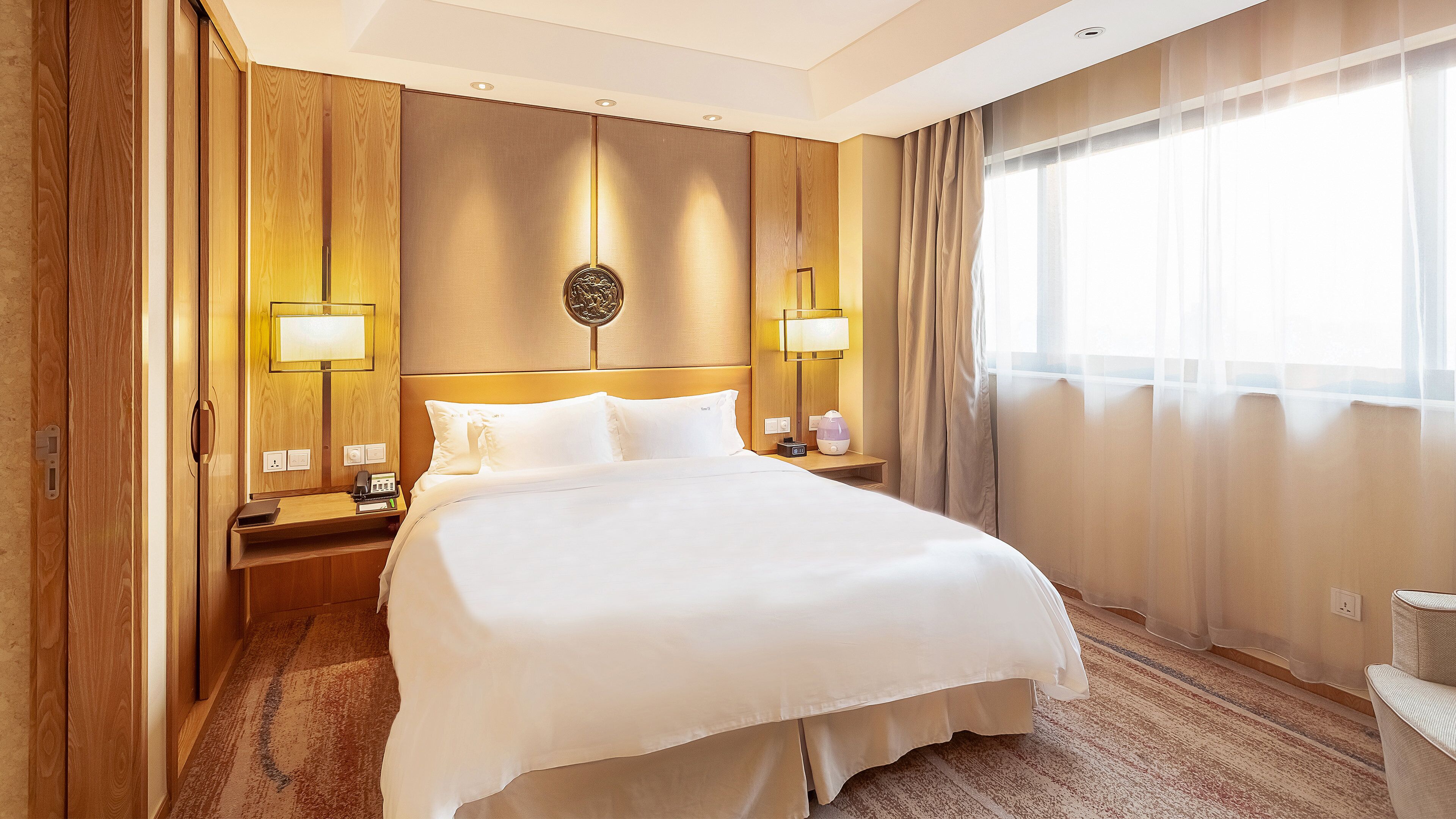 A hotel room with a large bed, wooden headboard, two bedside lamps, a nightstand, and a window with sheer curtains.