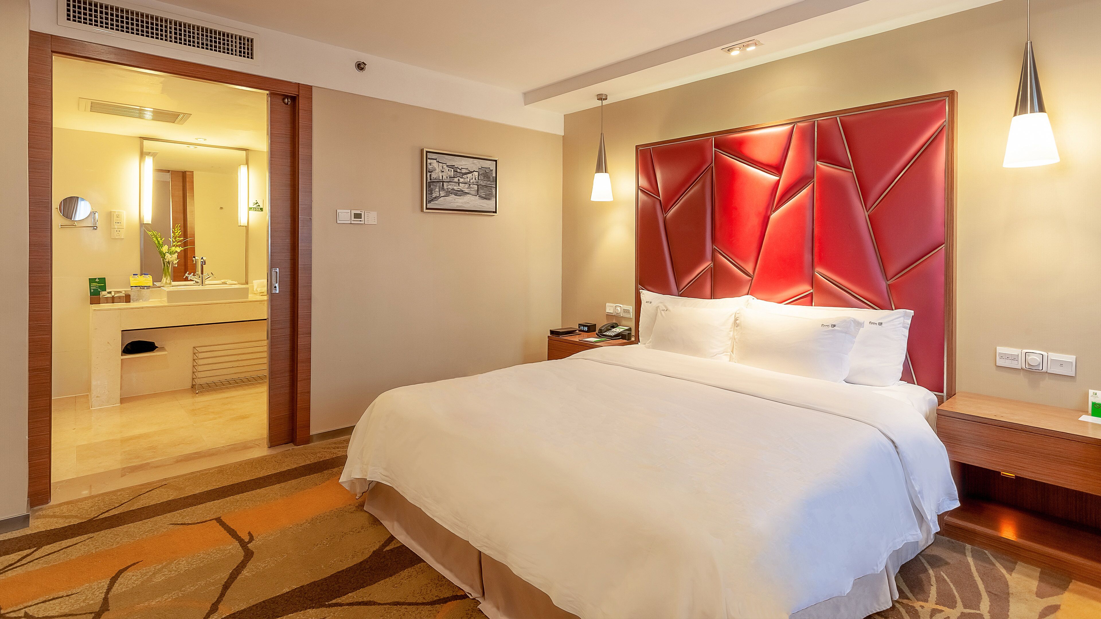 Ein Hotelzimmer mit einem Bett, einem Nachttisch, einem Spiegel, einem gerahmten Bild und einem roten Kopfteil.