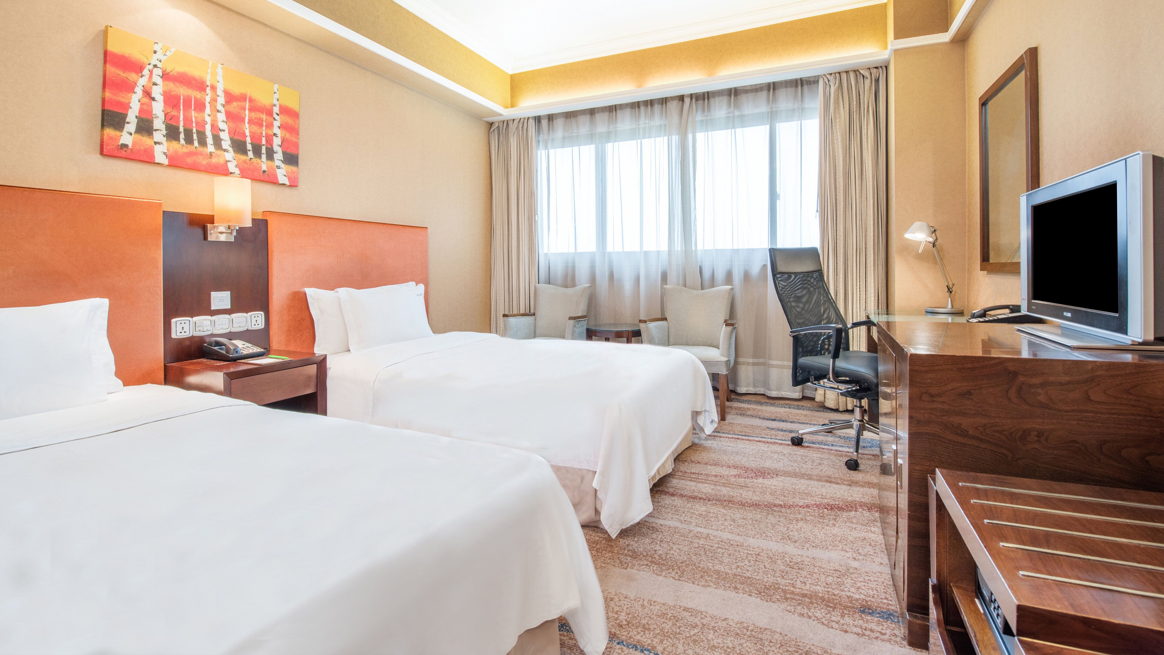 Habitación de hotel con dos camas, un escritorio con computadora, televisión y un ventanal con cortinas.