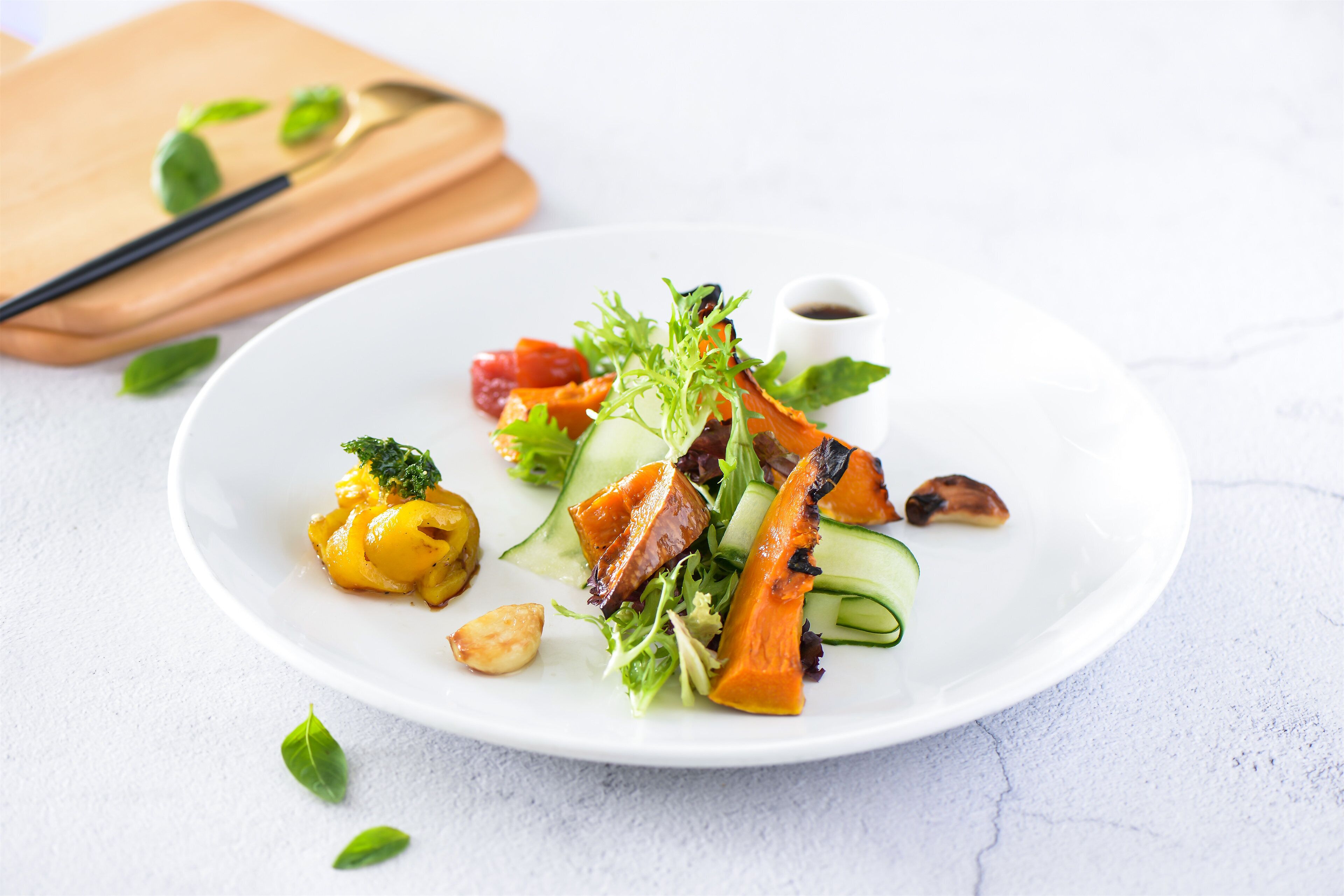 Een wit bord met een salade met gegrilde groenten, noten en een klein bakje saus.