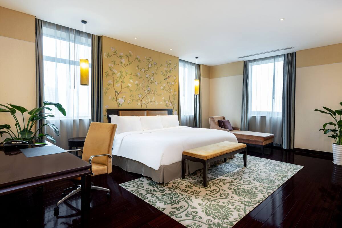 A hotel room with a large bed, a desk, a chair, a bench, and two potted plants.