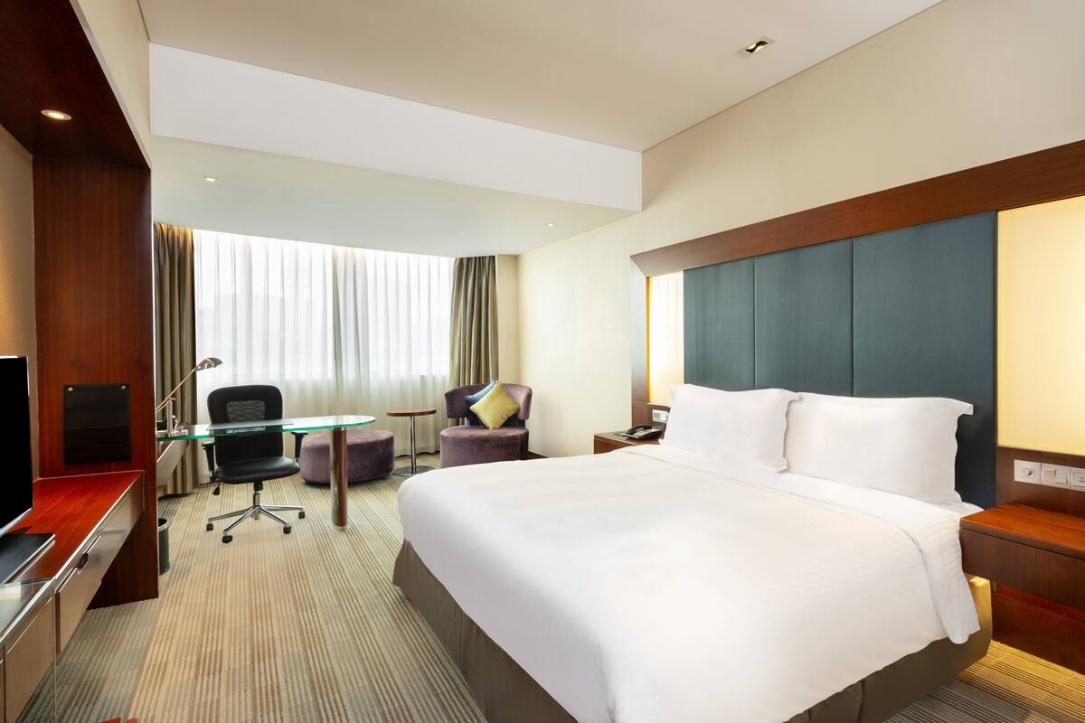 A hotel room with a large bed, a desk with a computer, a chair, a small table, and a window with curtains.