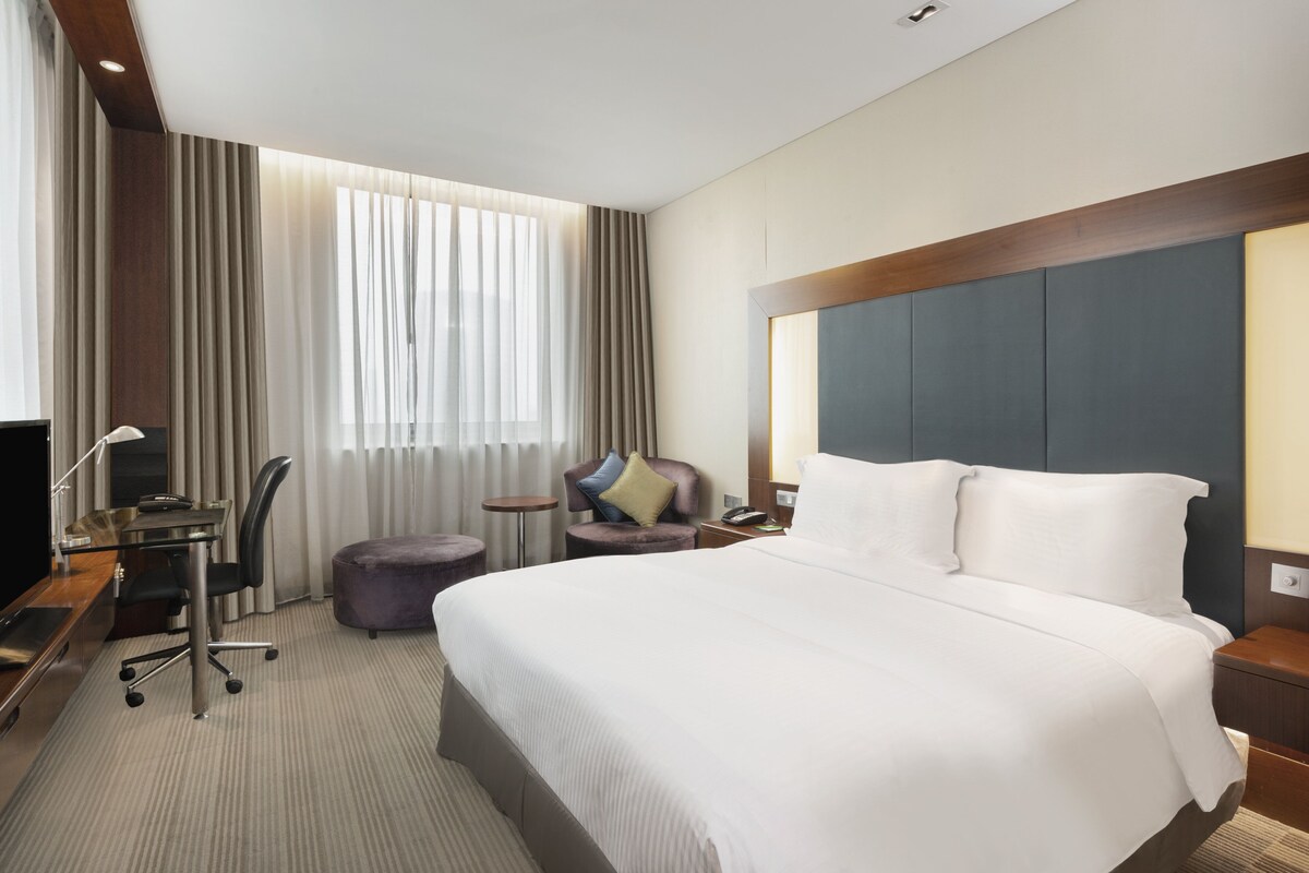 A hotel room with a large bed, a desk with a computer, a chair, a small table, and a window with curtains.