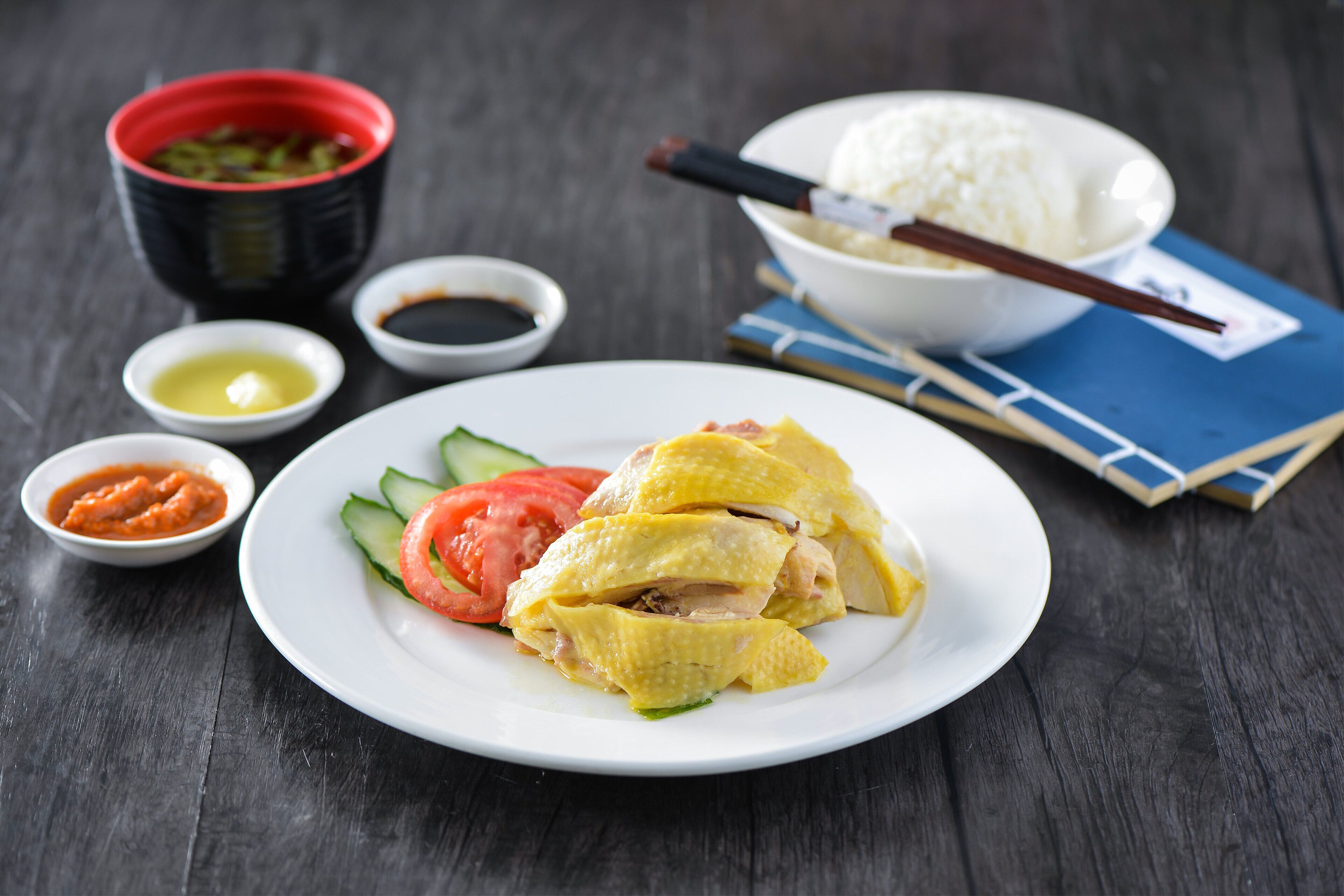 A plate of Hainanese chicken rice with a bowl of rice, chopsticks, and small bowls of sauces and condiments.