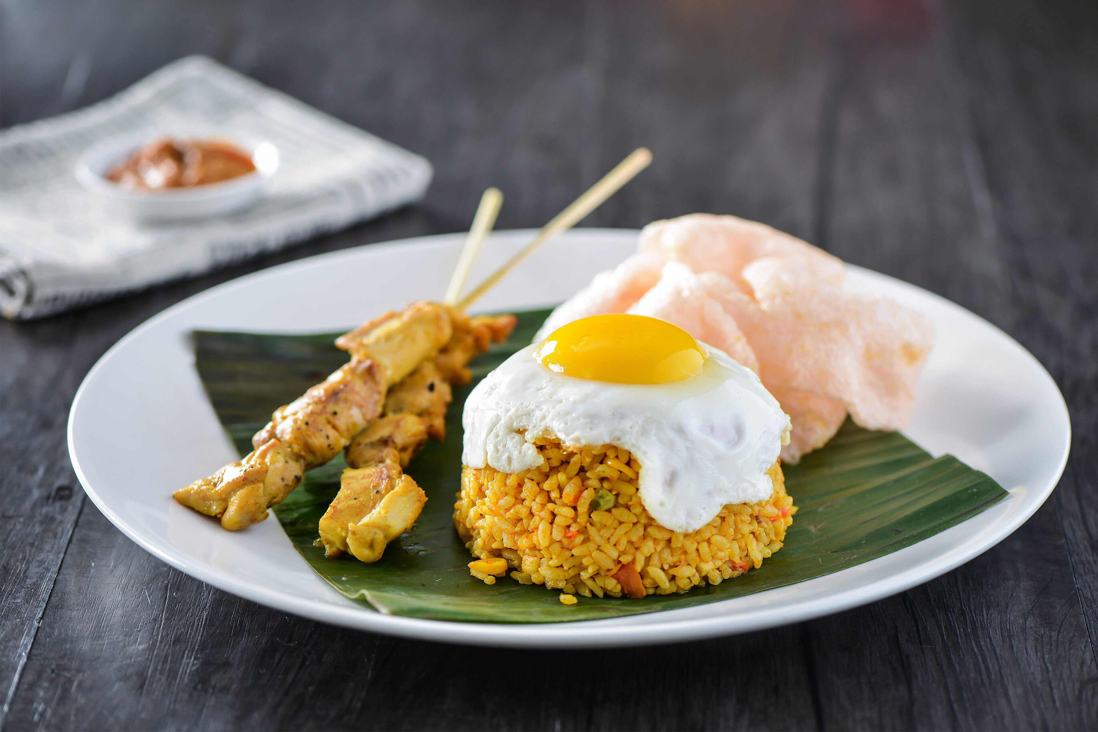 Une assiette de riz frit, surmontée d’un œuf au plat, accompagnée d’une brochette de poulet grillé et d’une tranche de pain.