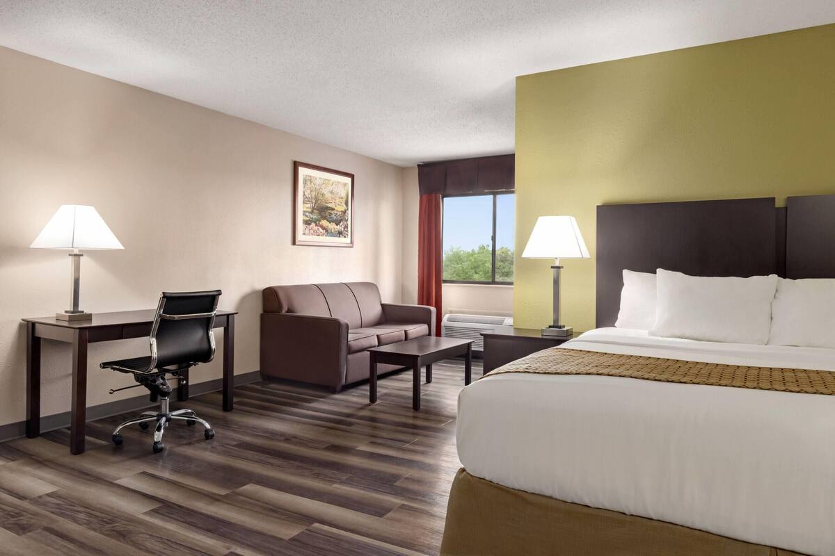 A hotel room with a bed, two chairs, a desk, a sofa, and a window with curtains.