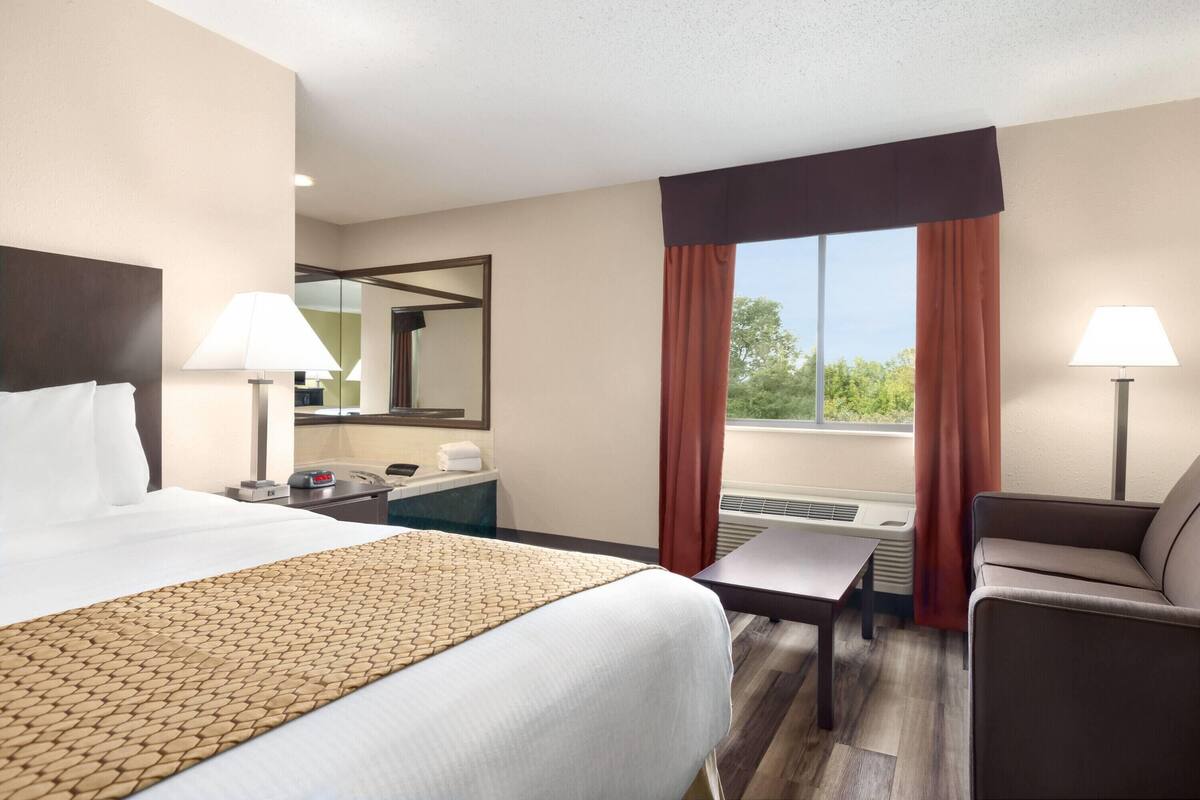 A hotel room with a bed, a desk, a chair, a window with curtains, and a mirror.