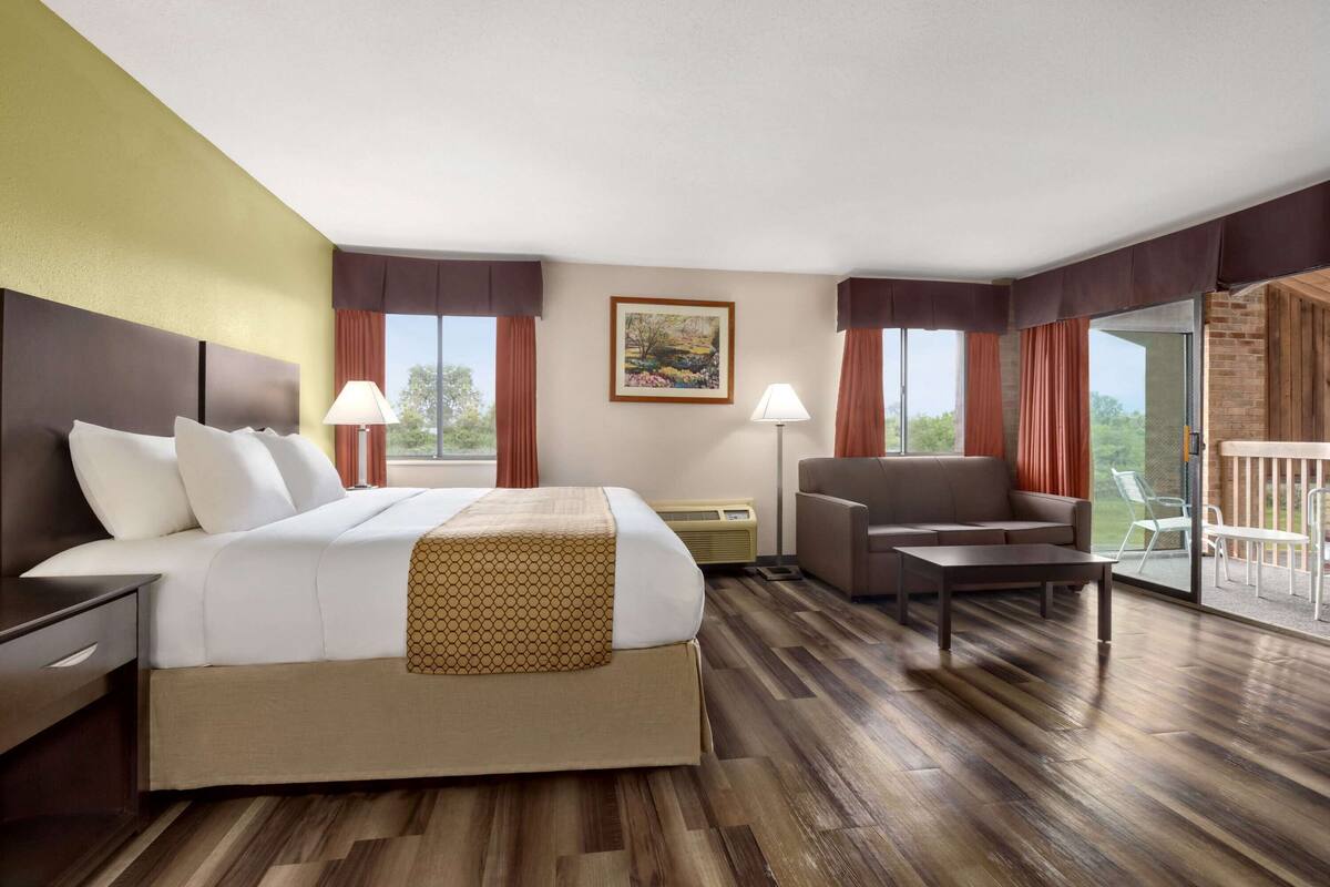 A hotel room with a large bed, a sofa, a small table, a chair, a painting on the wall, and a balcony with outdoor furniture.