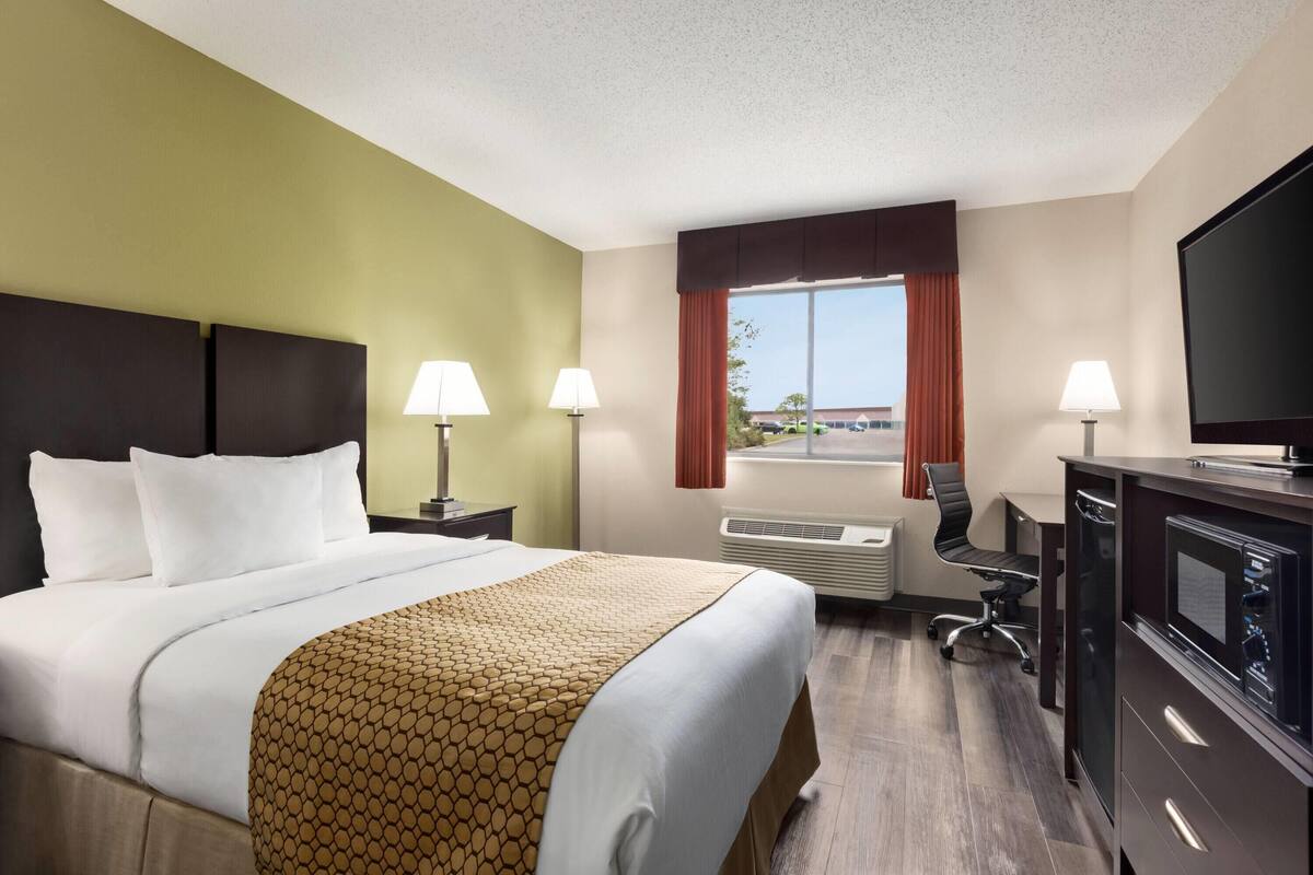 A hotel room with a bed, two bedside lamps, a desk with a chair, a television, and a window with curtains.