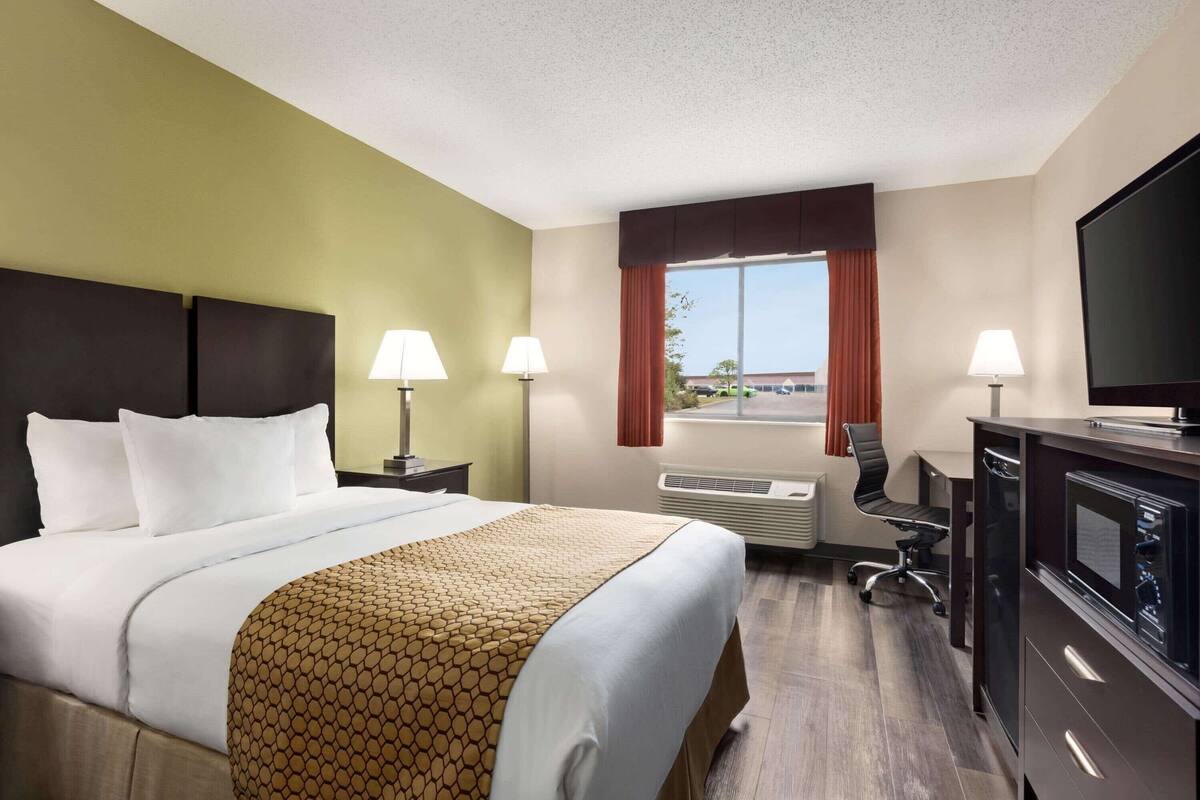 A hotel room with a bed, two bedside lamps, a desk with a chair, a television, and a window with curtains.