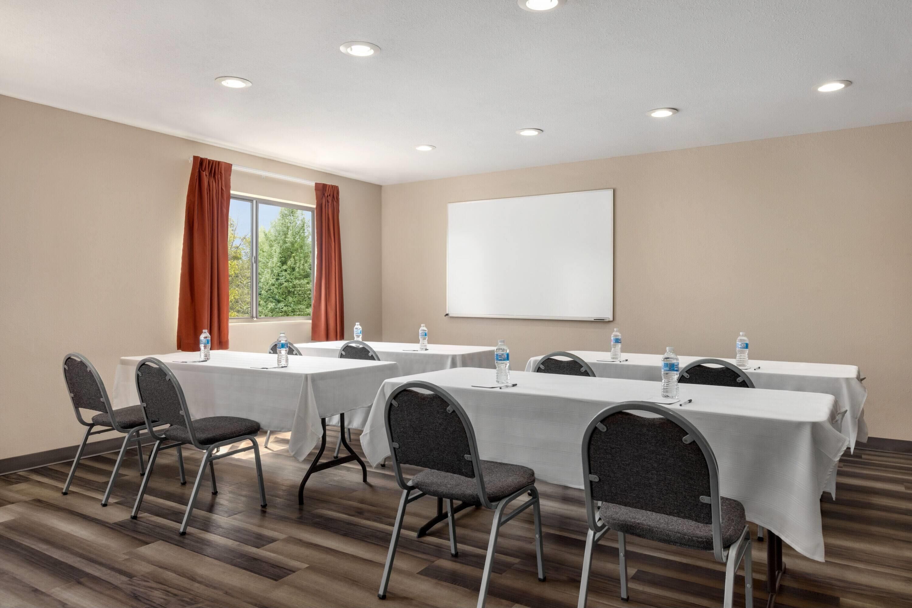 Un salón de conferencias con cuatro mesas, cada una con mantel blanco y botellas de agua. El salón tiene piso de madera, paredes color beige y una pizarra grande.