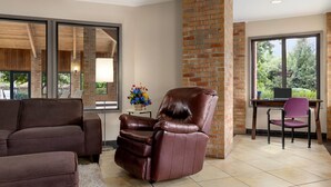A modern living room with a leather recliner, a brown sofa, and a purple chair. There is a brick accent wall, a large window with a view of greenery, and a small table with a vase of flowers. - Baymont by Wyndham Monroe (Monroe)