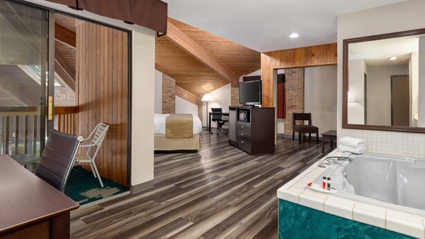 A hotel room with a wooden floor, a large window, a bed, a desk with a computer, a chair, a small table, and a bathtub. - Baymont by Wyndham Monroe (Monroe)